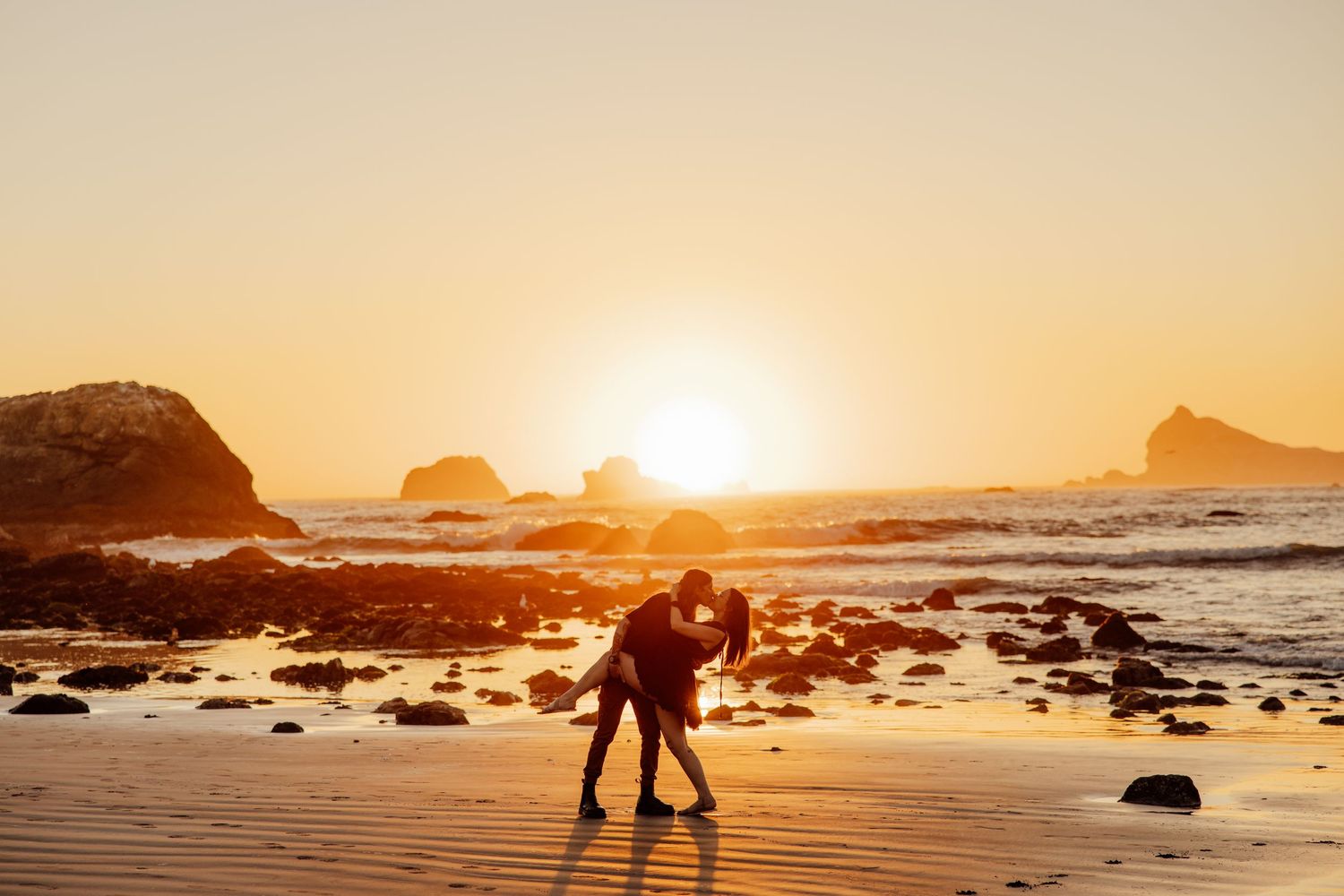 Dramatic ocean sunset with waves crashing against rocky coastline and silhouetted figures on beach.