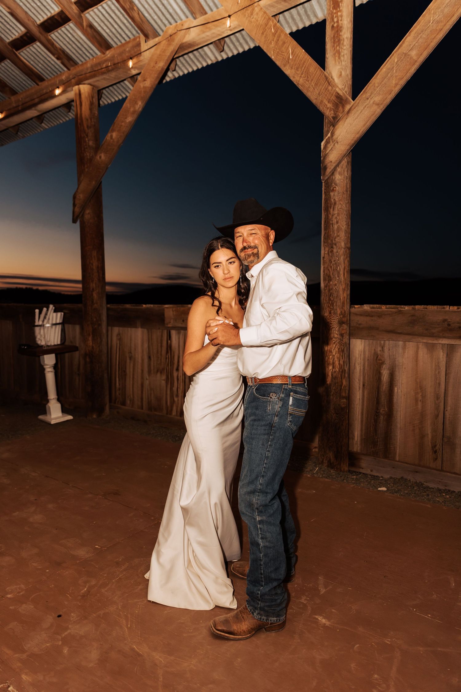 Newlyweds sharing first dance in rustic barn with wooden beams at sunset.