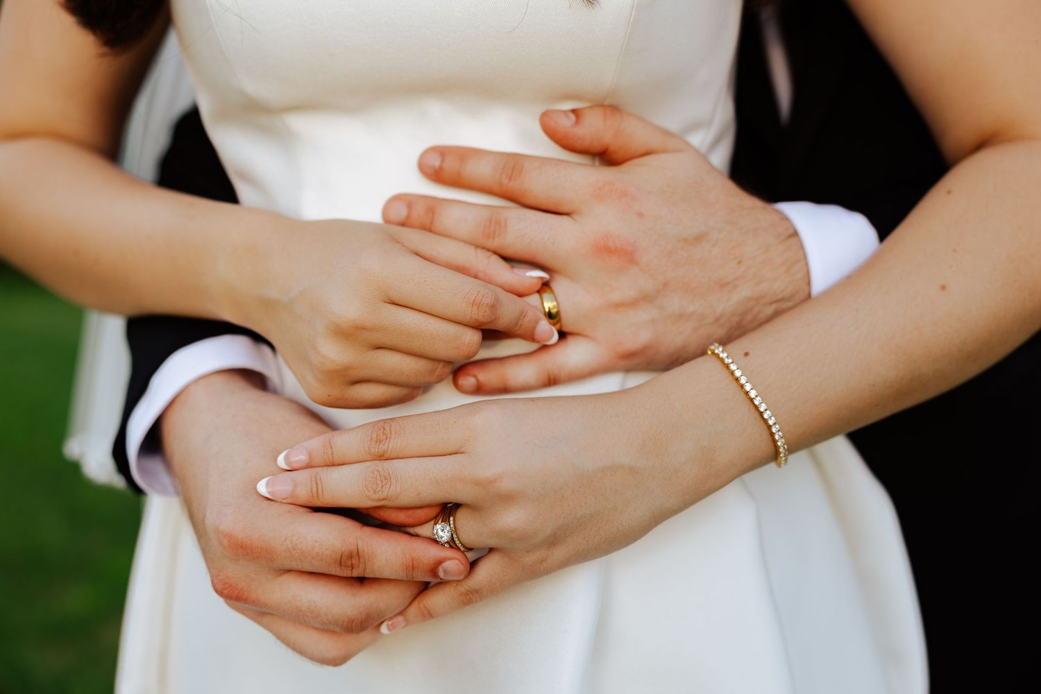 Newlywed couple displaying wedding rings on clasped hands, bride in white dress with French manicure and gold bracelet.