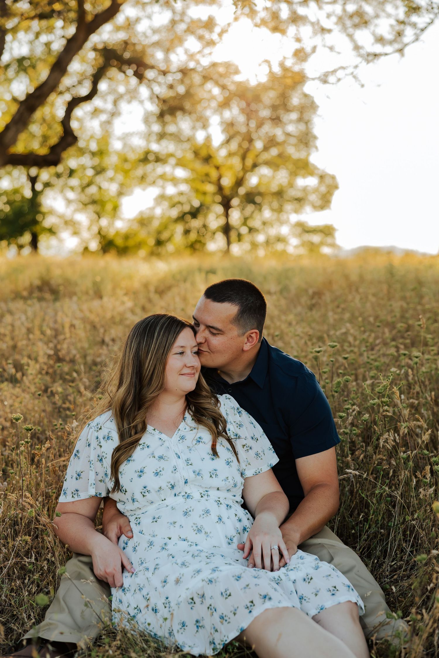 Couple sitting in tall golden grass field at sunset, man kissing womans forehead.