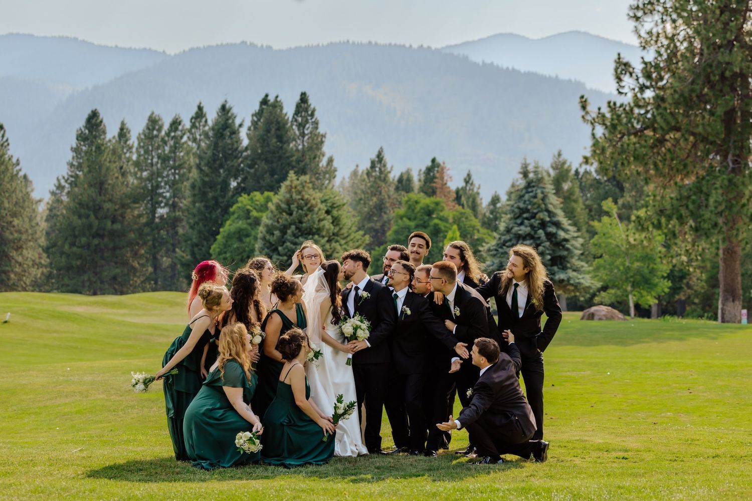 Wedding party in emerald green and black attire posing on a lush golf course with mountain views.