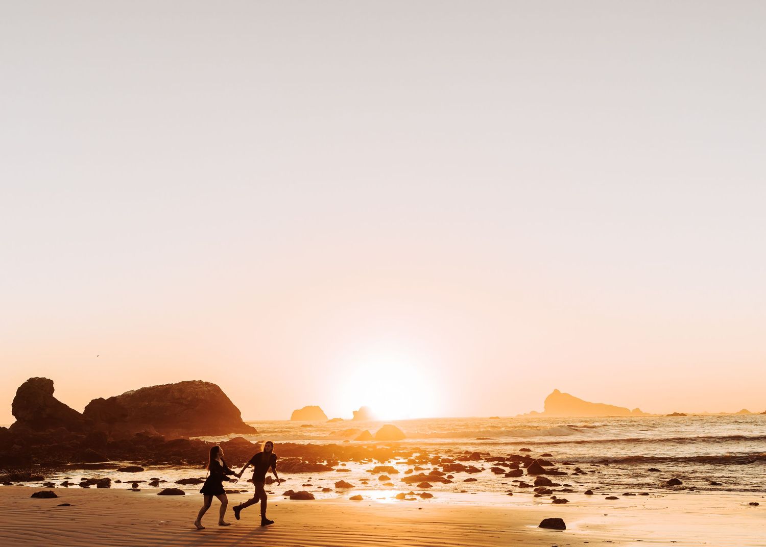 Two figures walking along beach at sunset with rocky coastline and ocean waves.