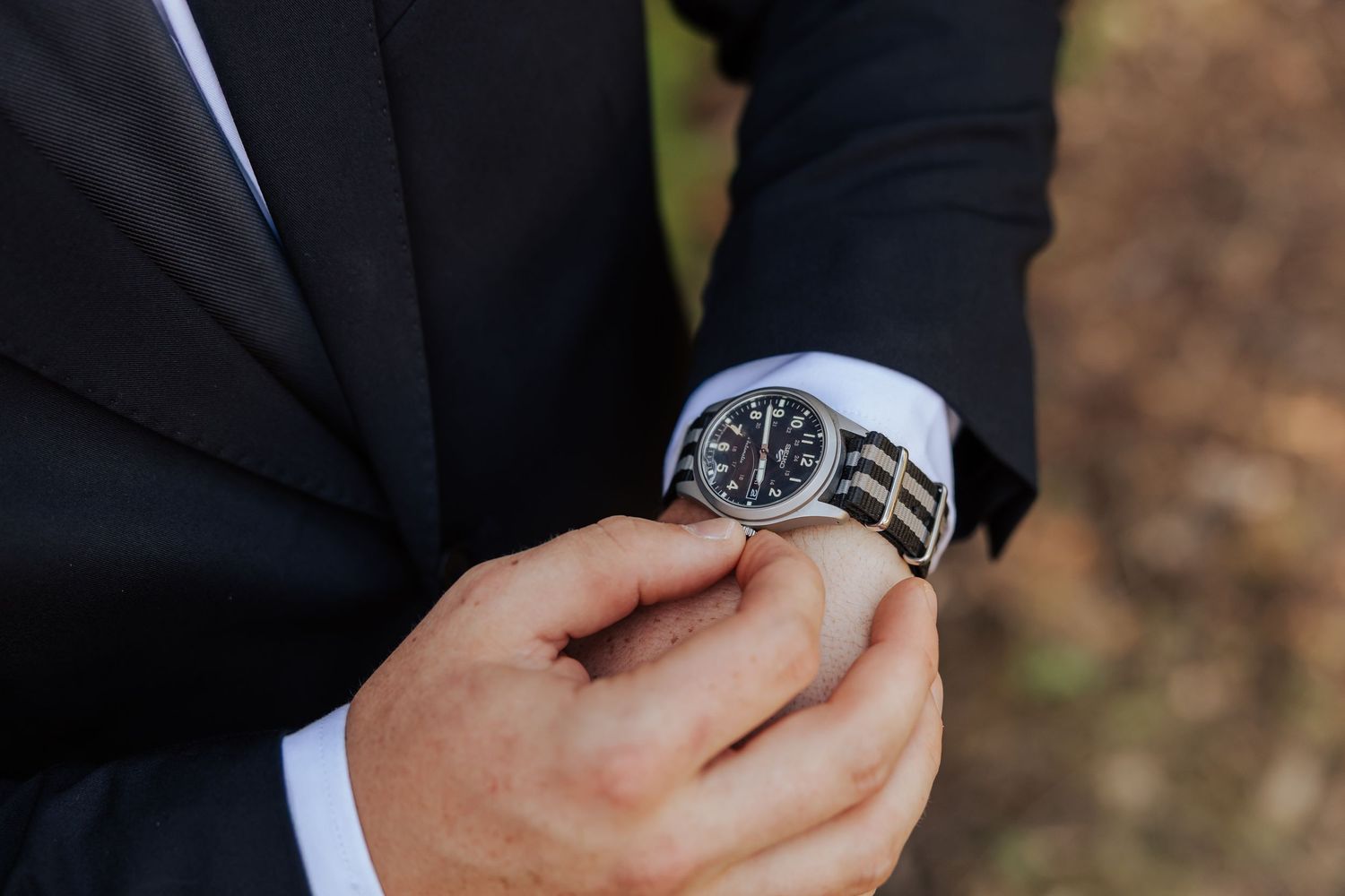 Groom adjusts his classic black watch on his wrist while wearing a sharp dark suit on his wedding day.