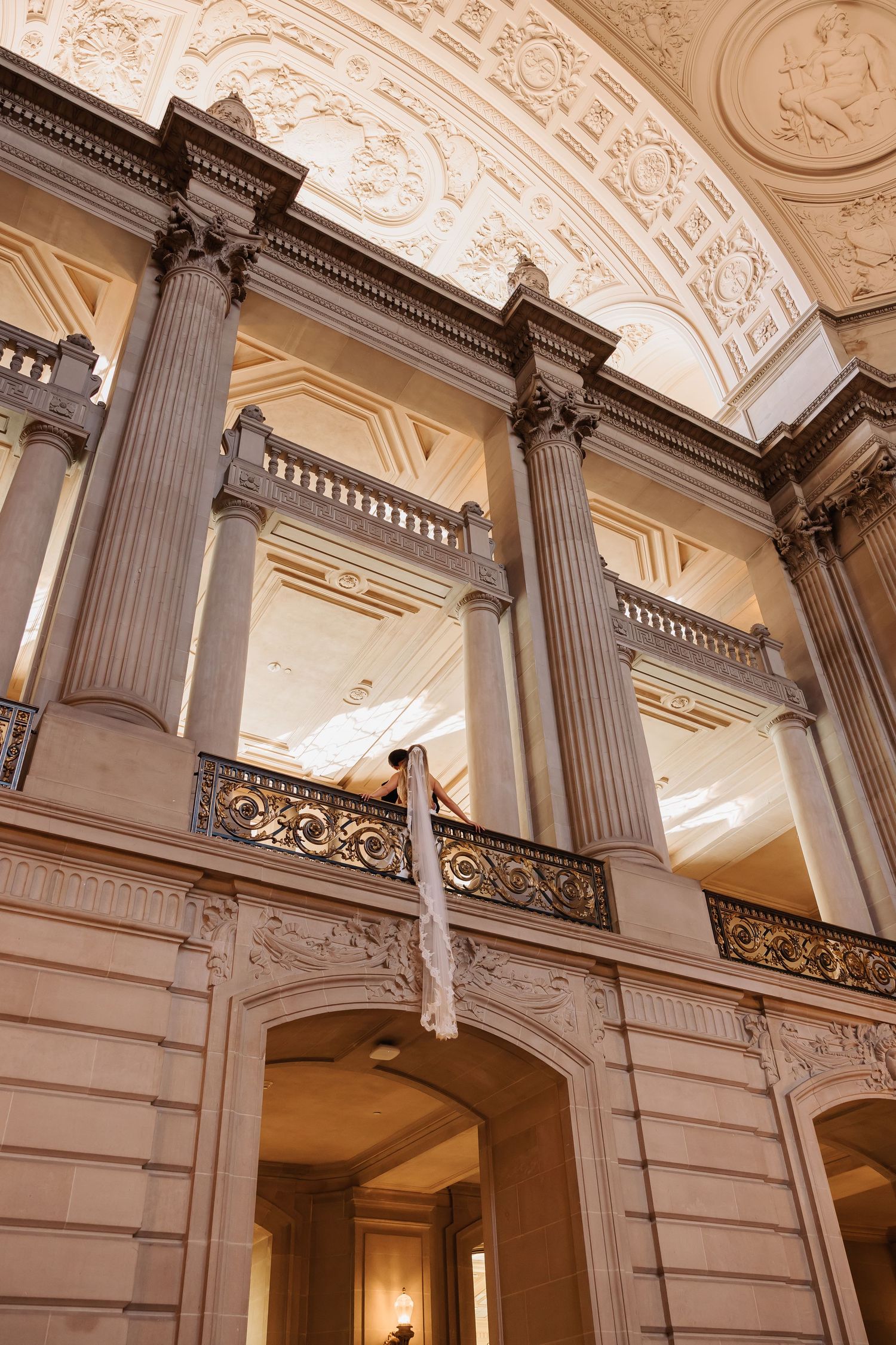Bride in wedding dress stands on ornate balcony of grand neoclassical building with tall columns and gilded railings.