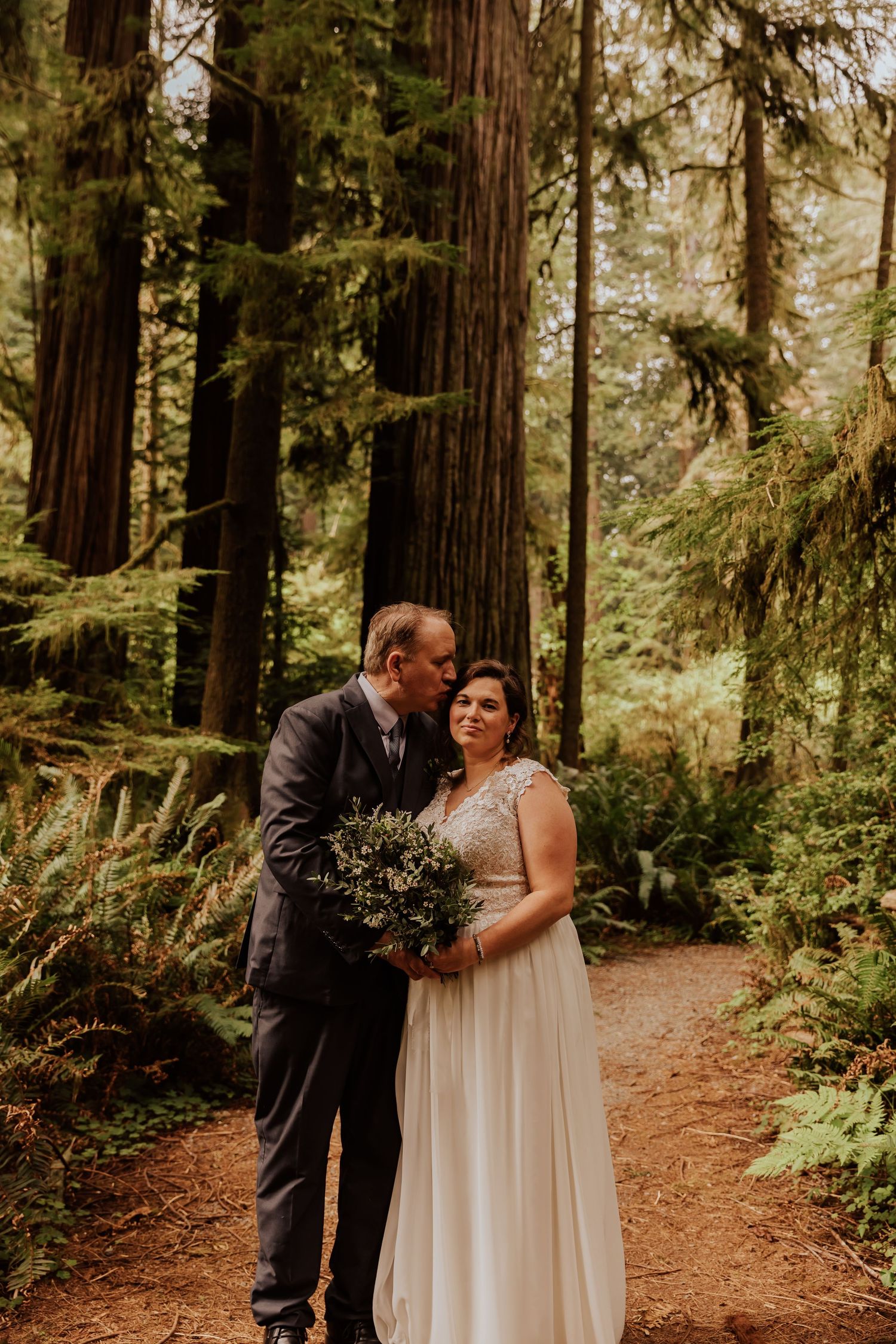 A romantic wedding moment captured on a forest trail surrounded by towering redwood trees.