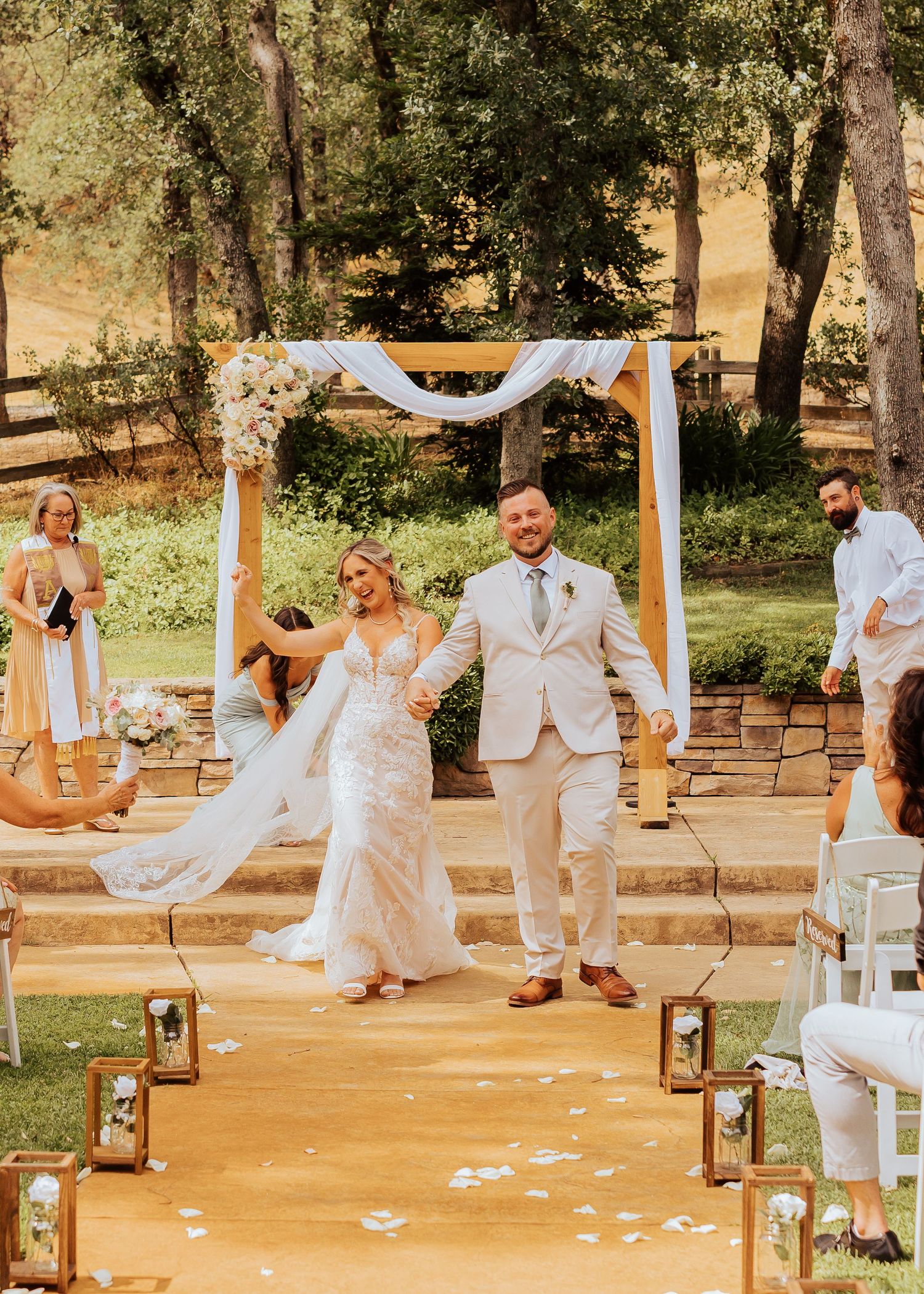 Wedding party in white and sage green attire gathered around a decorated outdoor ceremony space.