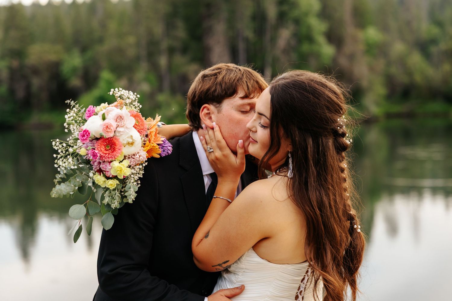 Bride and groom sharing a kiss outdoors near a lake, bride holding a colorful bouquet of flowers.