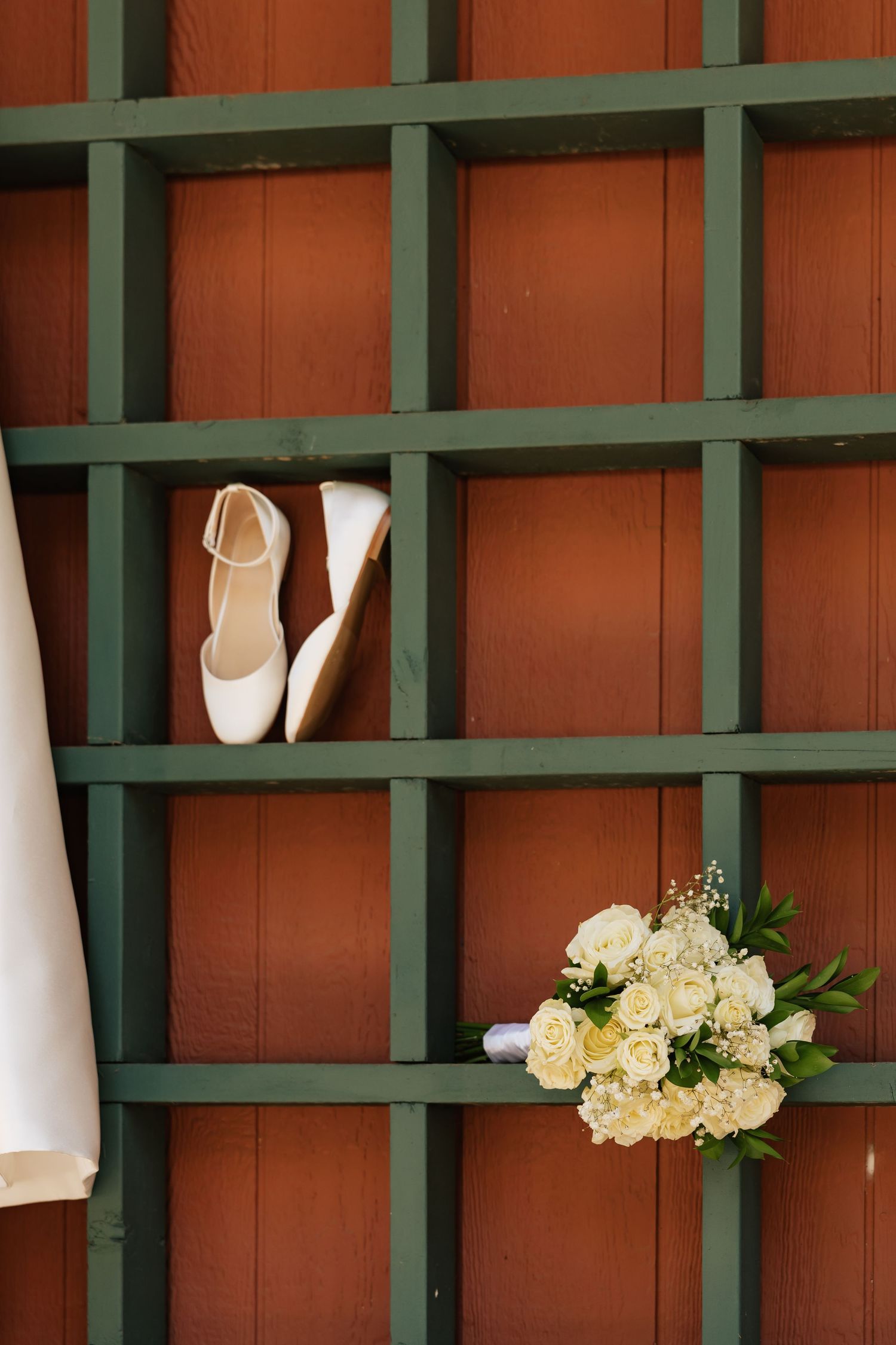 White bridal heels and a bouquet of white roses displayed on a green lattice wooden wall for a wedding.