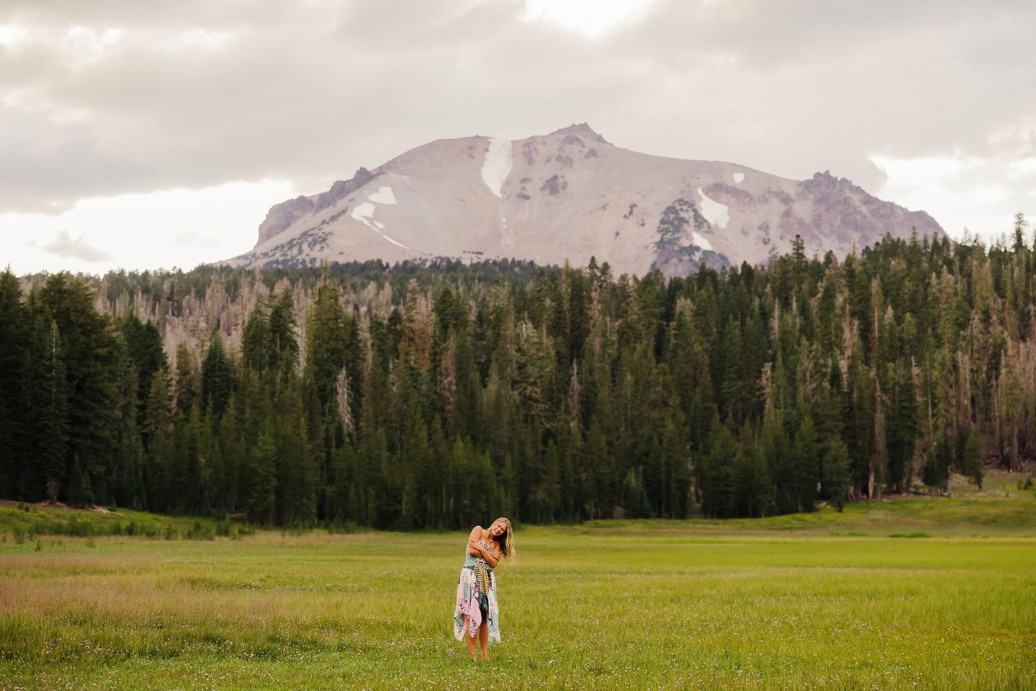 Couple embracing in a green meadow with tall pine trees and a snow-capped mountain peak under cloudy skies.