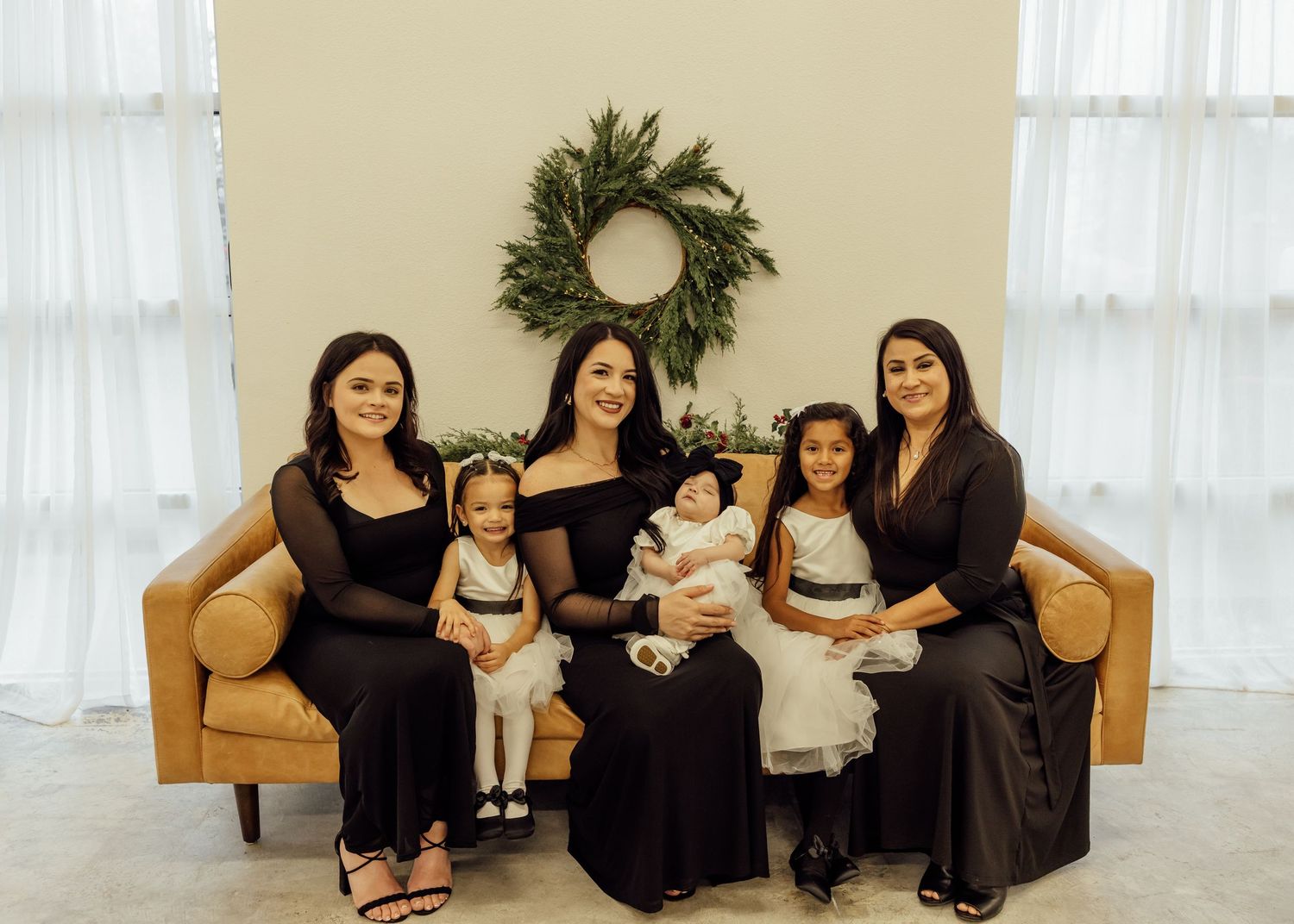 A group wearing black dresses sits together on a beige couch in a bright studio setting with white curtains.