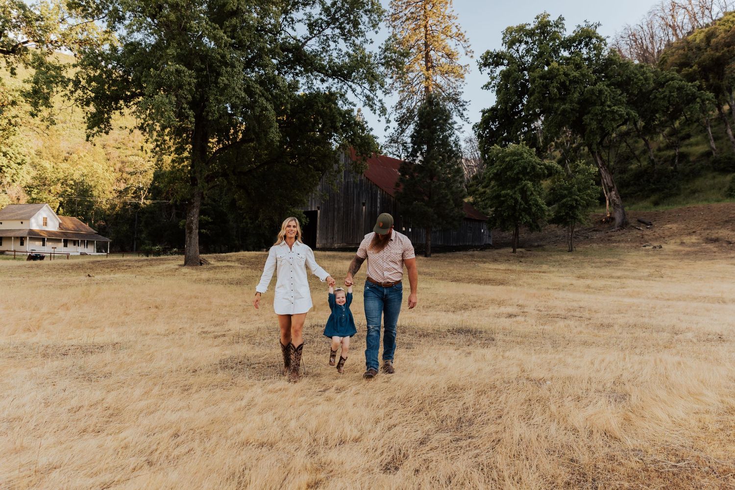 Three people stroll through a sun-drenched field with trees and a barn in the background.