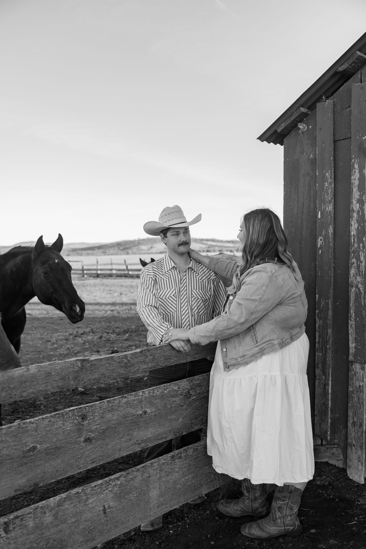 A couple stands by a wooden fence with horses visible in the rural background.