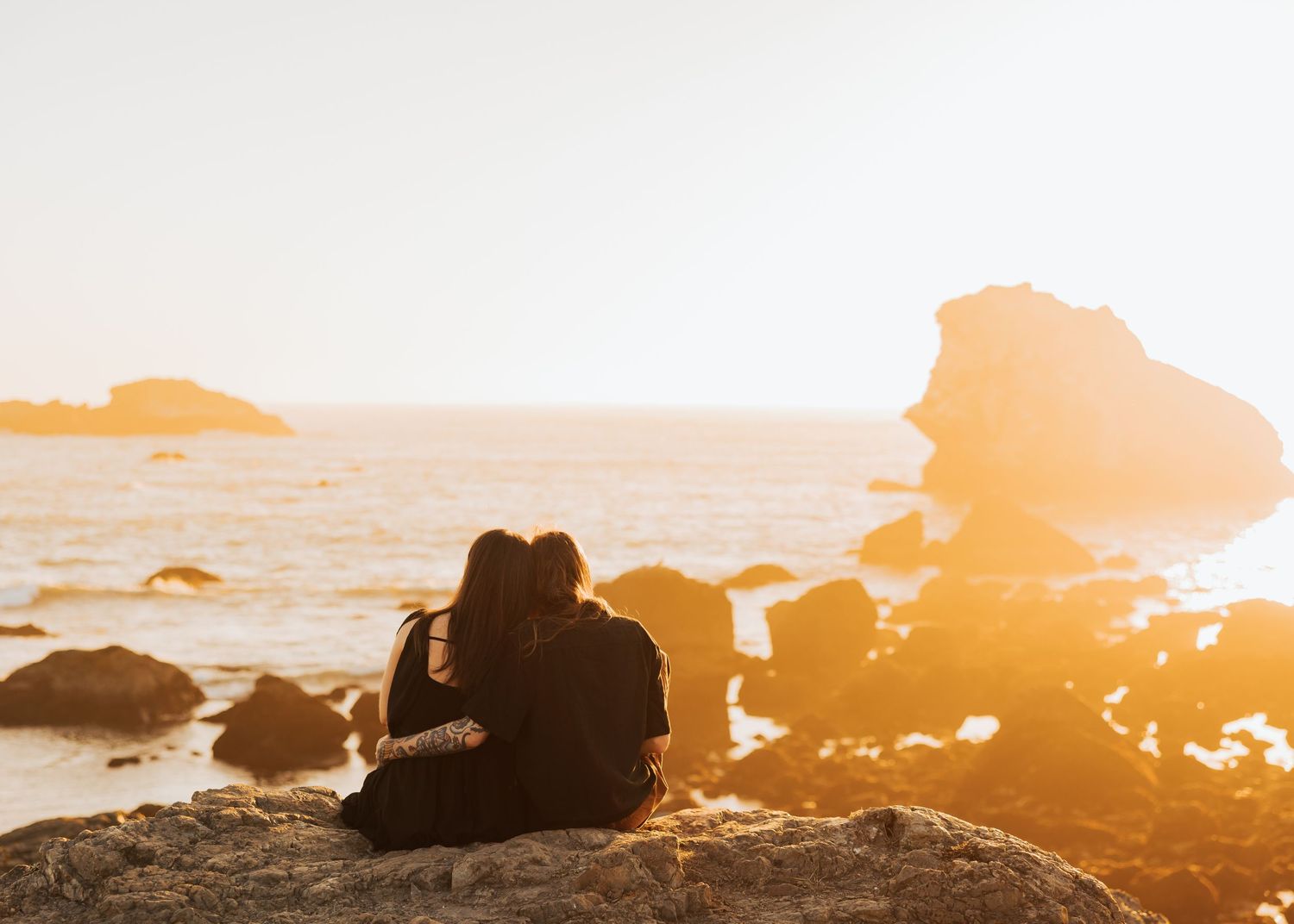 Silhouetted figures sit together on rocky shore watching the sunset over the ocean.