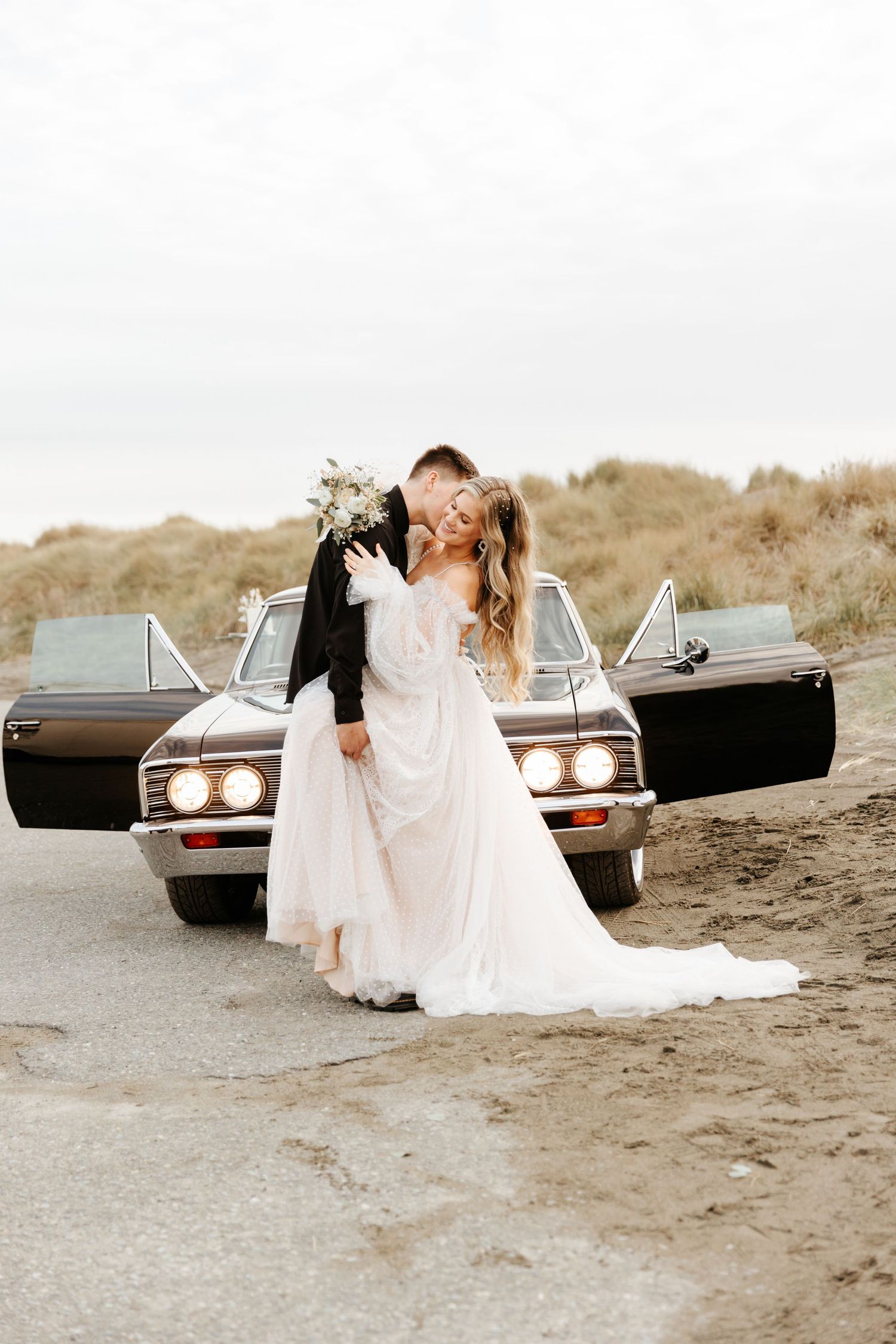 Newlyweds share a kiss in front of vintage car with doors open on beach.