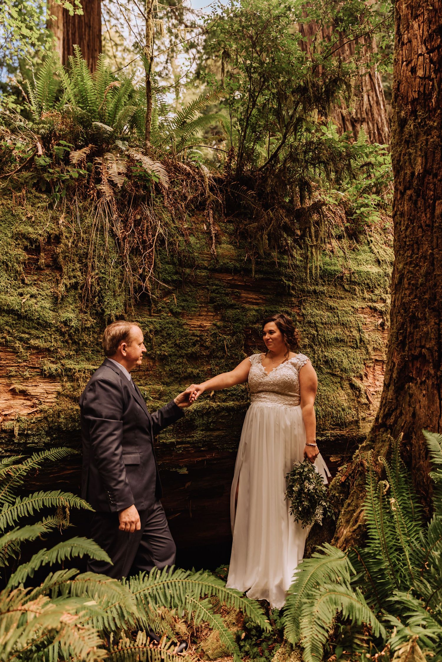 Playful wedding couple interaction among ferns and redwoods on a forest path.