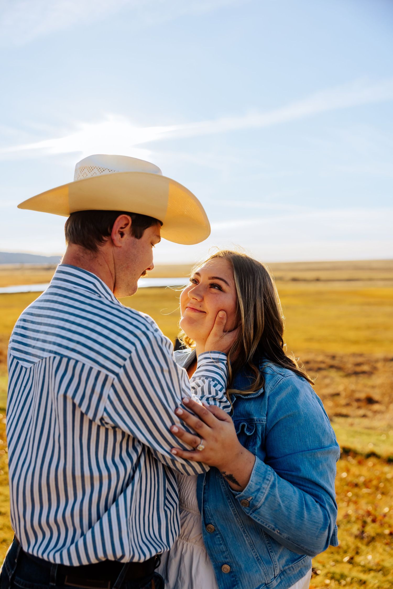 A romantic couple embraces in a golden field at sunset wearing western attire and cowboy hats.