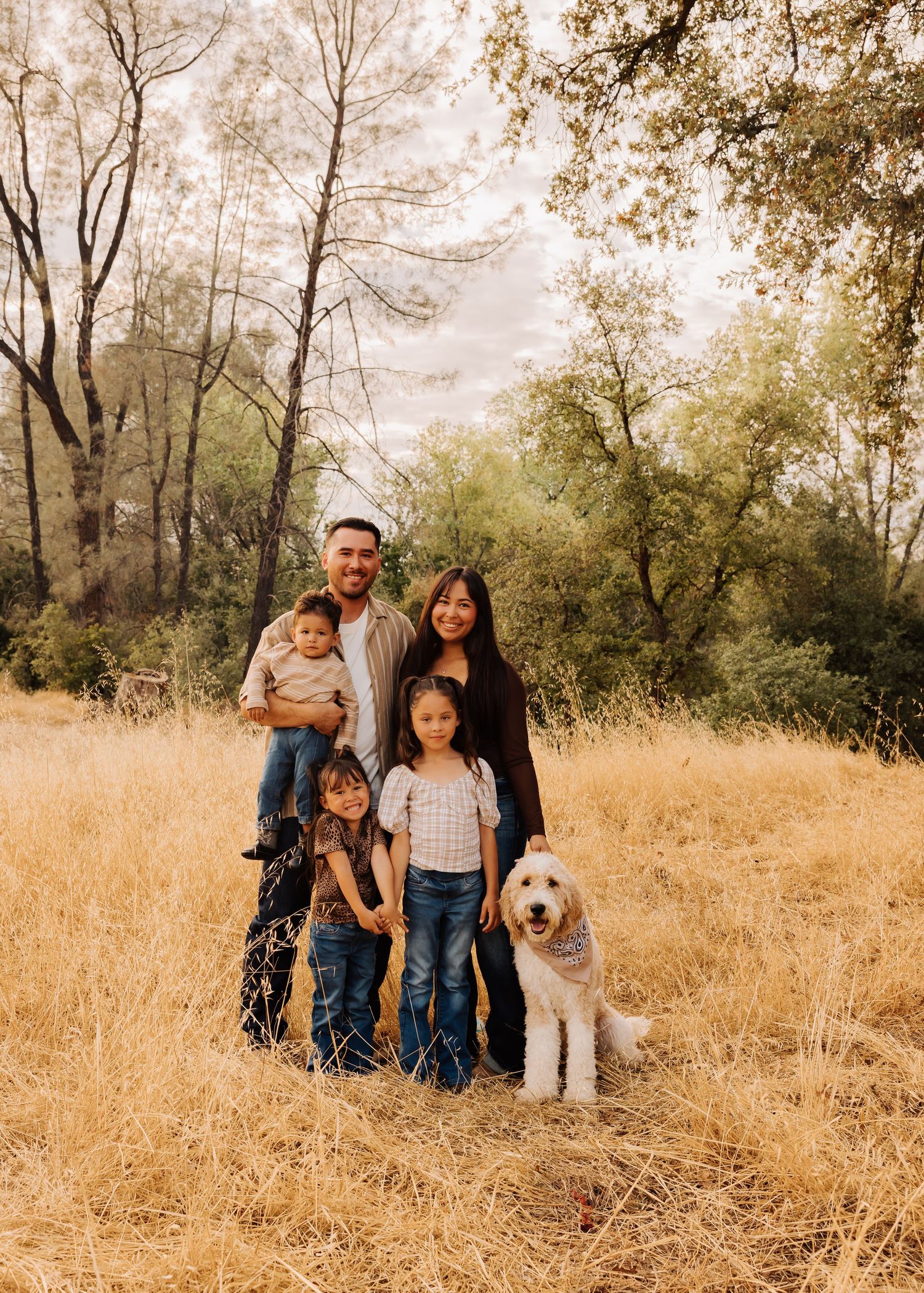 Family and dog pose together in golden field with trees in background during autumn.