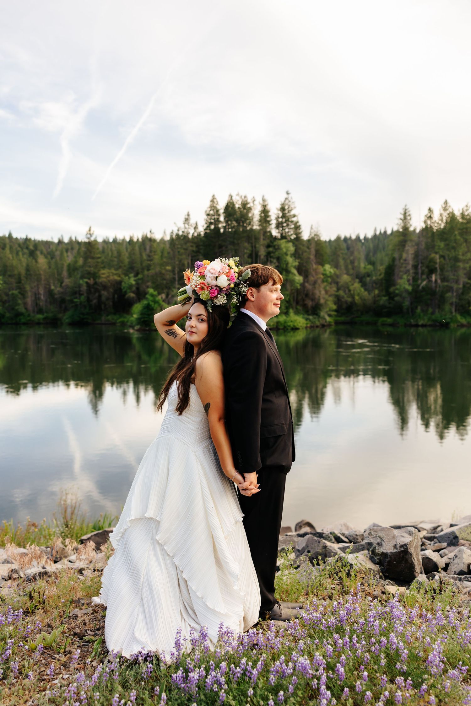 Bride and groom pose by a serene forest lake, bride in flowing white gown with floral crown holding colorful bouquet.