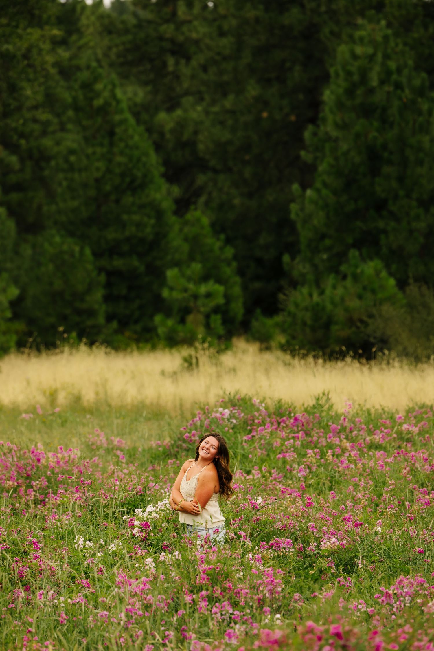 Woman standing in a wildflower meadow with pink blooms, surrounded by lush green forest trees.