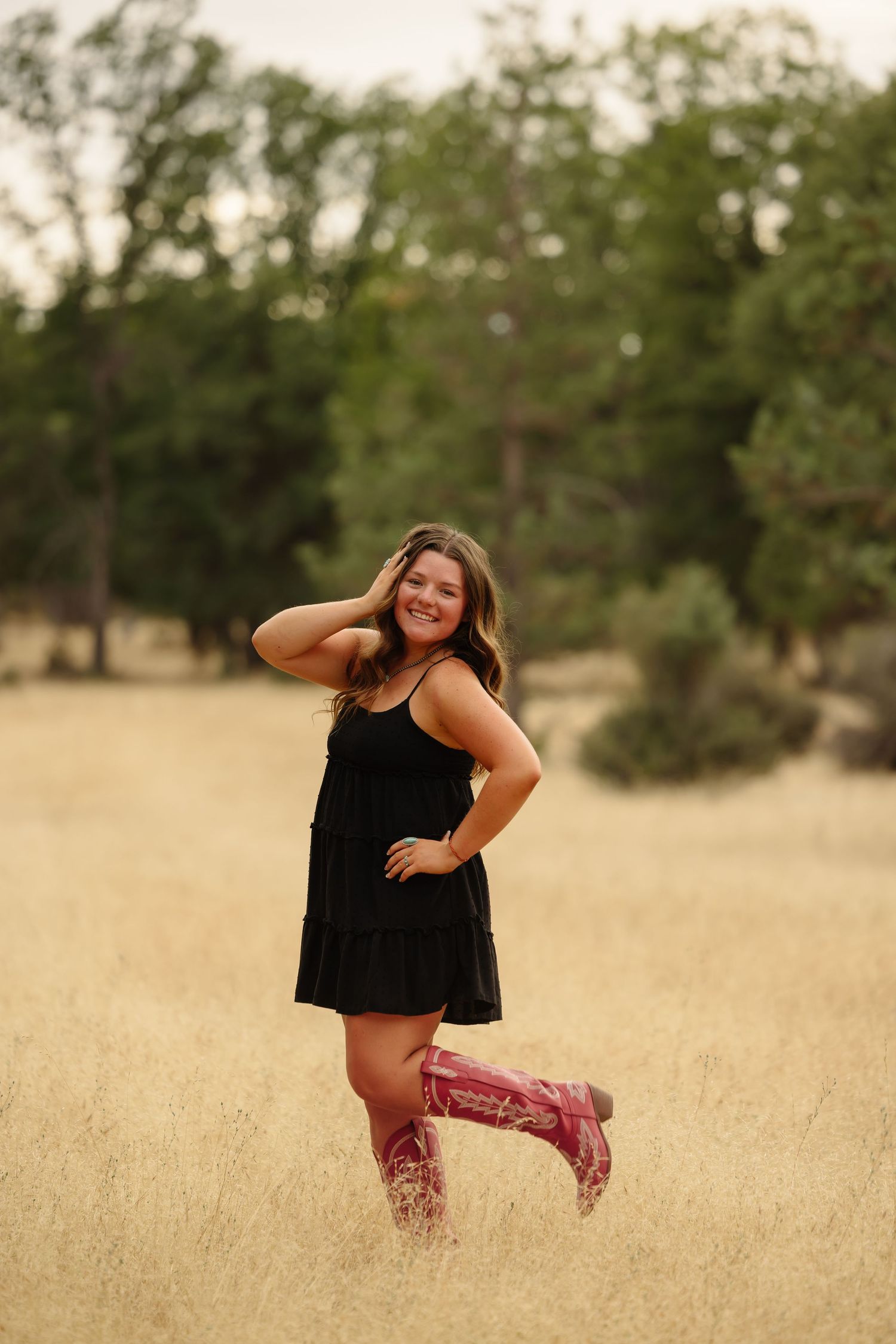 Young woman in black dress and pink cowboy boots posing in golden field with trees in background.