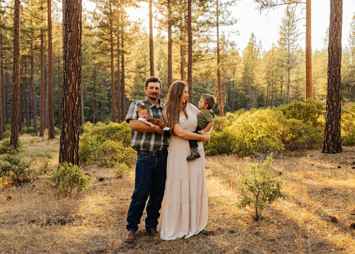 Family photoshoot in casual attire against sunlit pine forest backdrop.