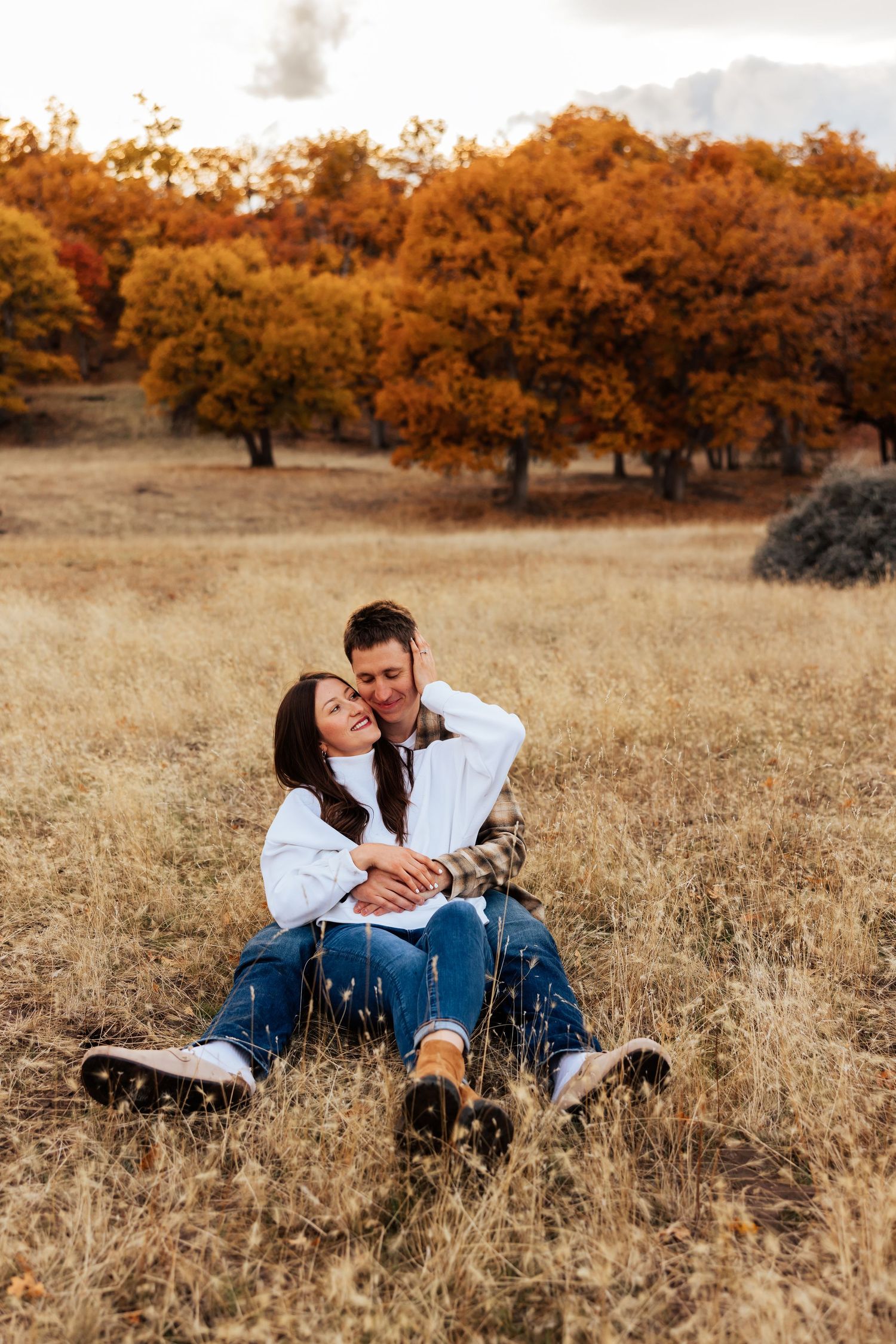 Couple sharing tender moments while sitting in dry grass with autumn trees behind them.