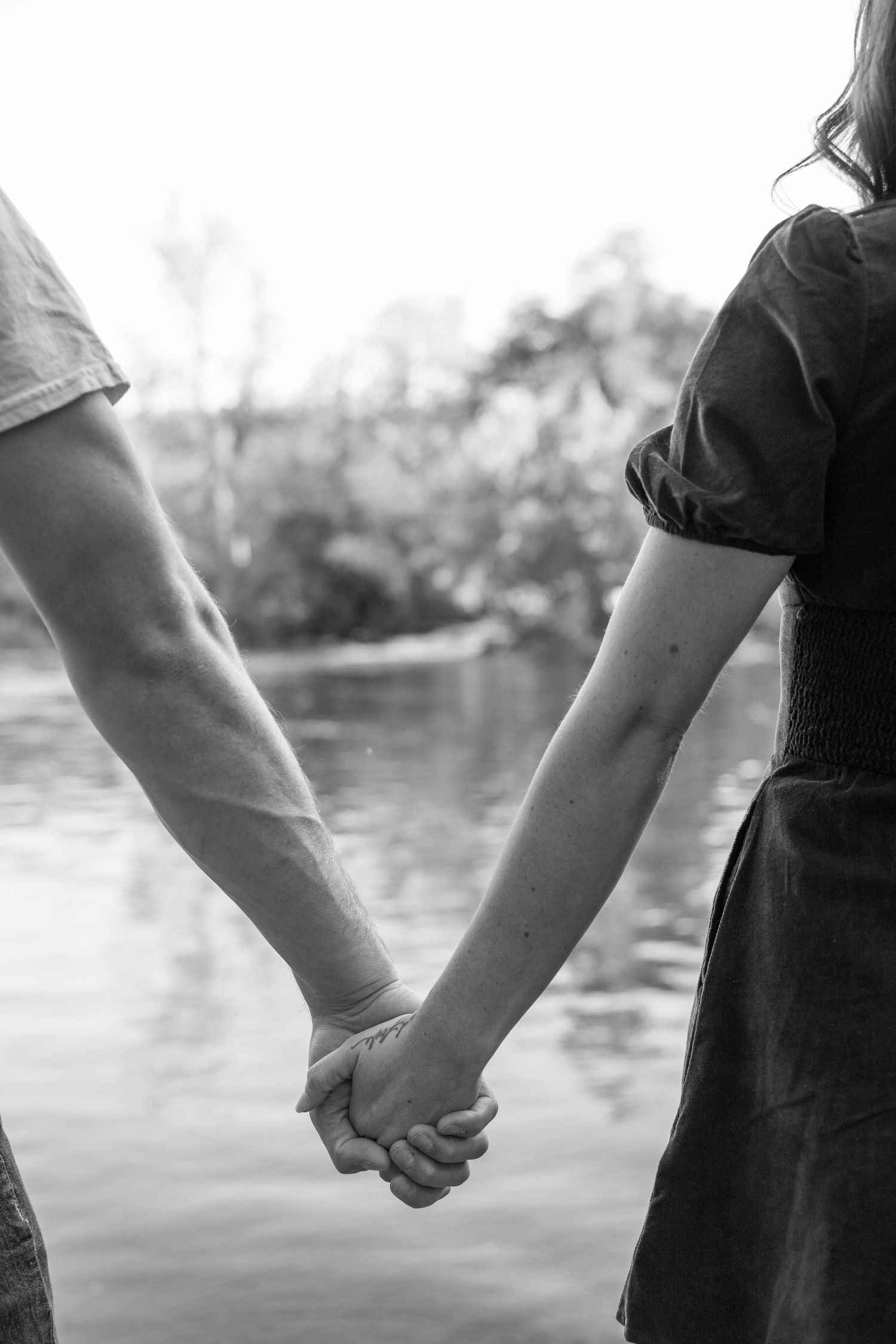 Black and white close-up of two people holding hands by a lake.