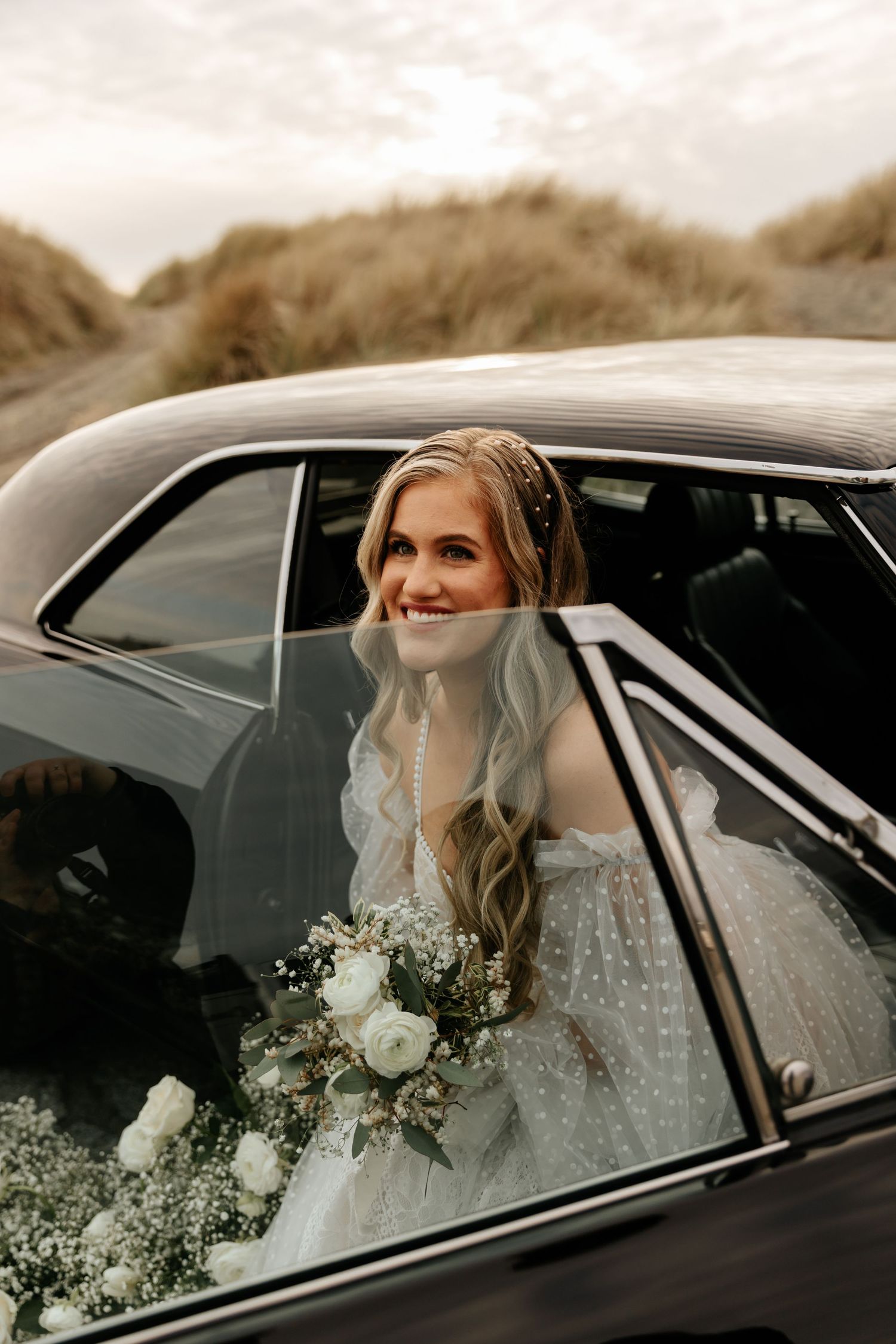 Bride in lace dress sitting in classic black car with white floral bouquet.