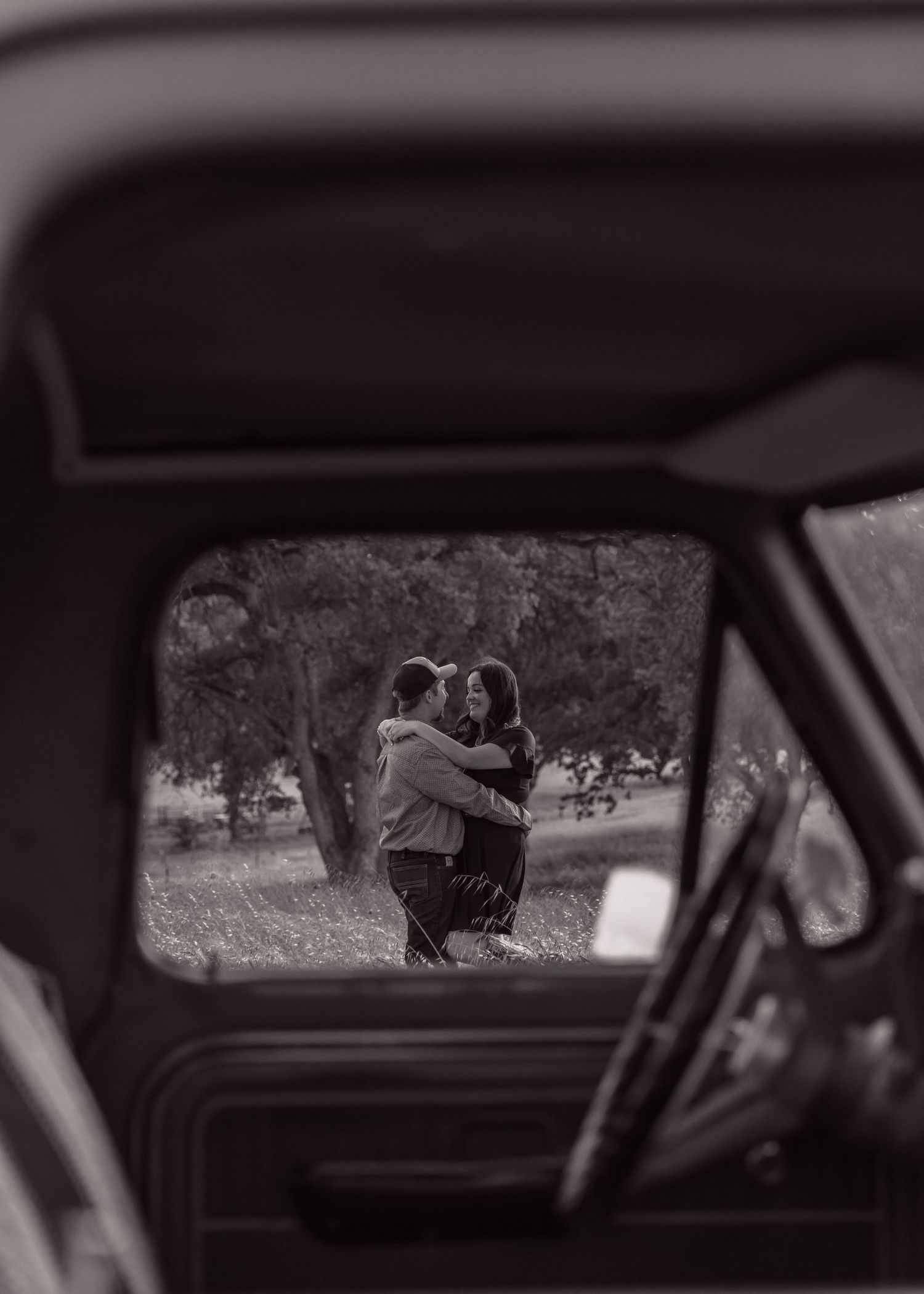 A romantic couple embracing is viewed through a car window frame in black and white photography.