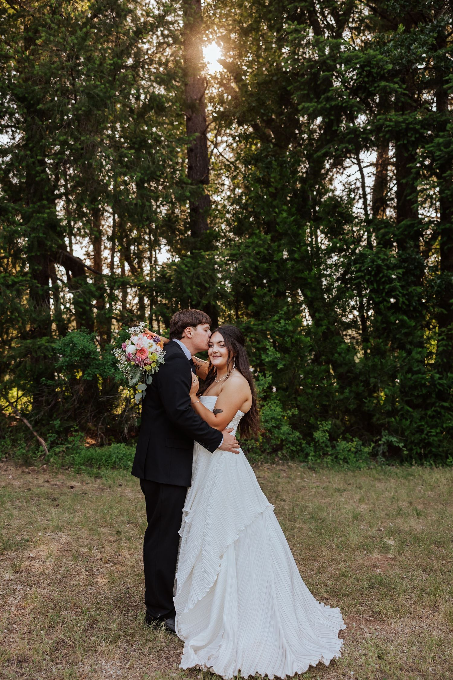 Bride and groom embrace at golden hour in a lush forest, groom kissing bride's temple tenderly.