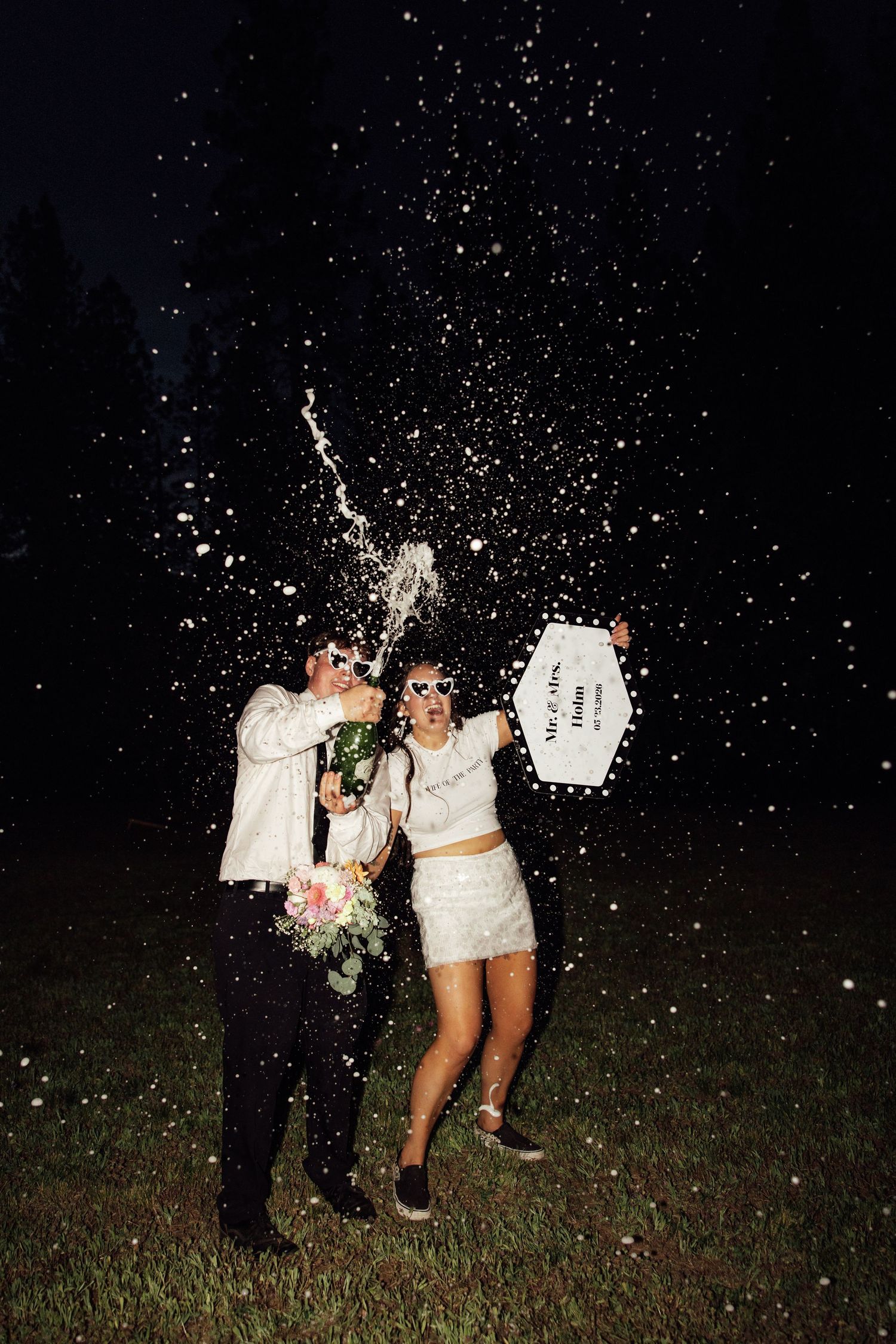 Joyful newlyweds celebrate with champagne spray at night, bride in white mini dress holding sign, groom in suit.