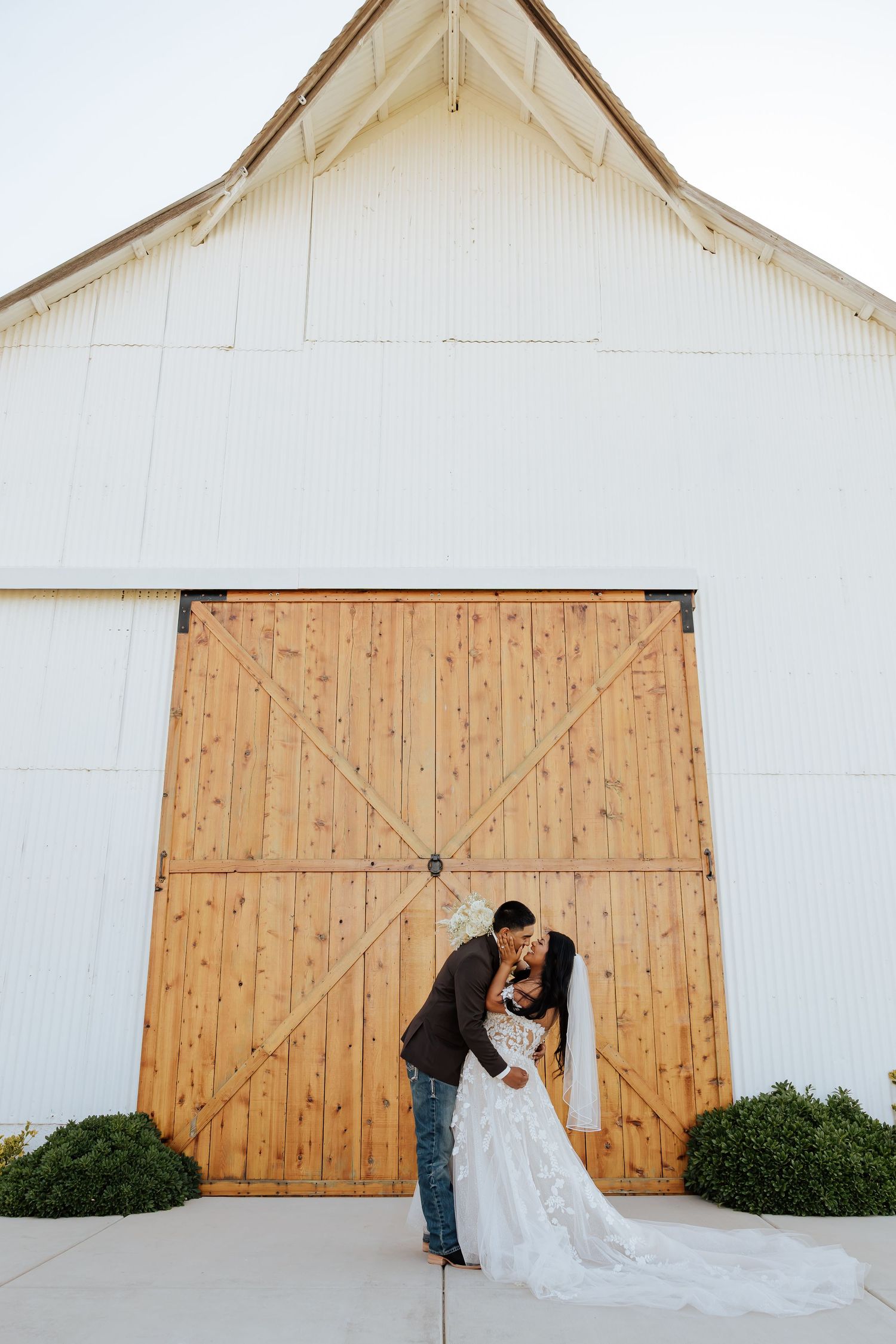 Bride and groom kissing in front of large wooden barn doors at a rustic white barn wedding venue.