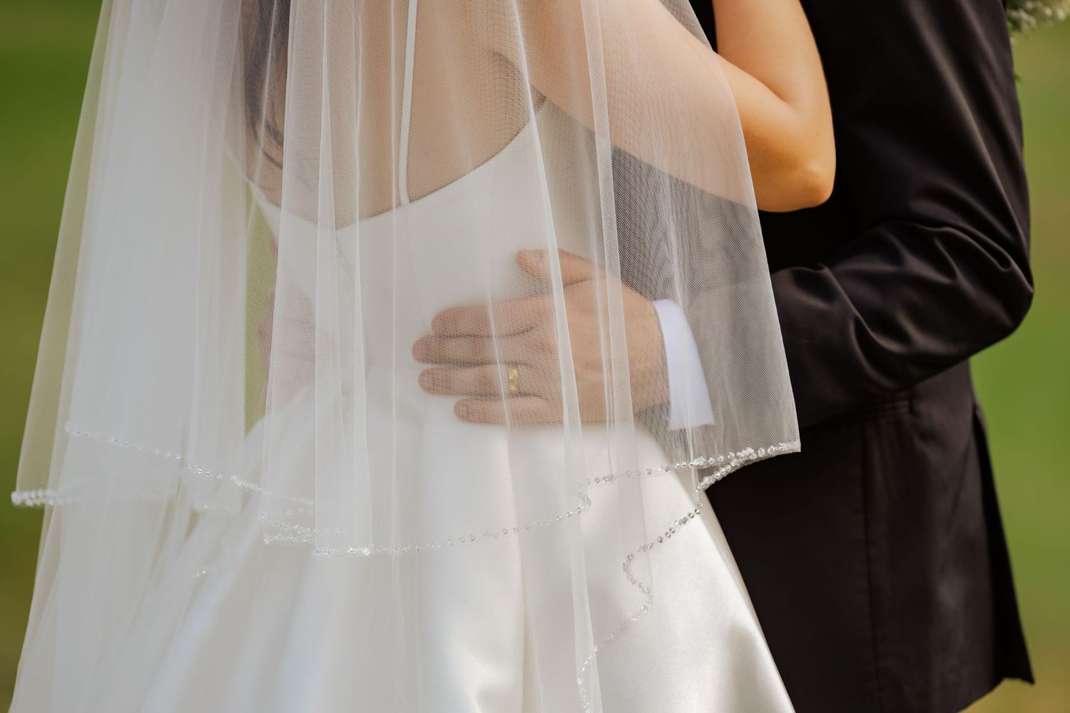 Bride and groom embracing, showcasing white wedding dress with veil and groom's black suit at outdoor ceremony.