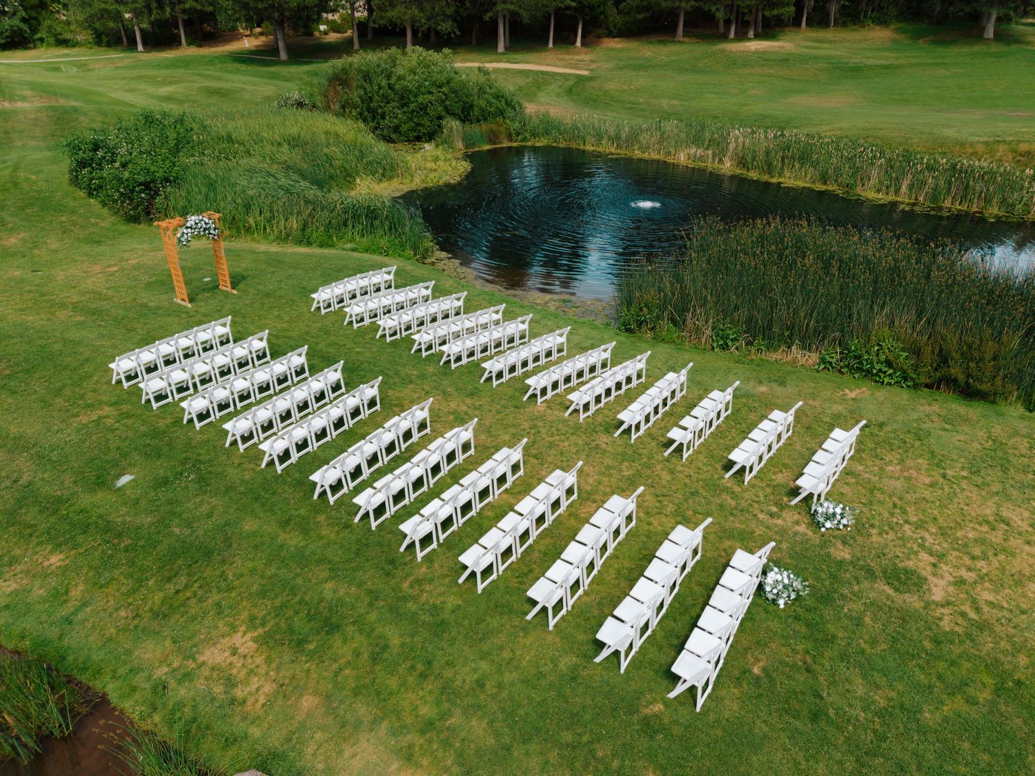 Aerial view of white chairs arranged in rows on green grass beside a pond for an outdoor wedding ceremony.