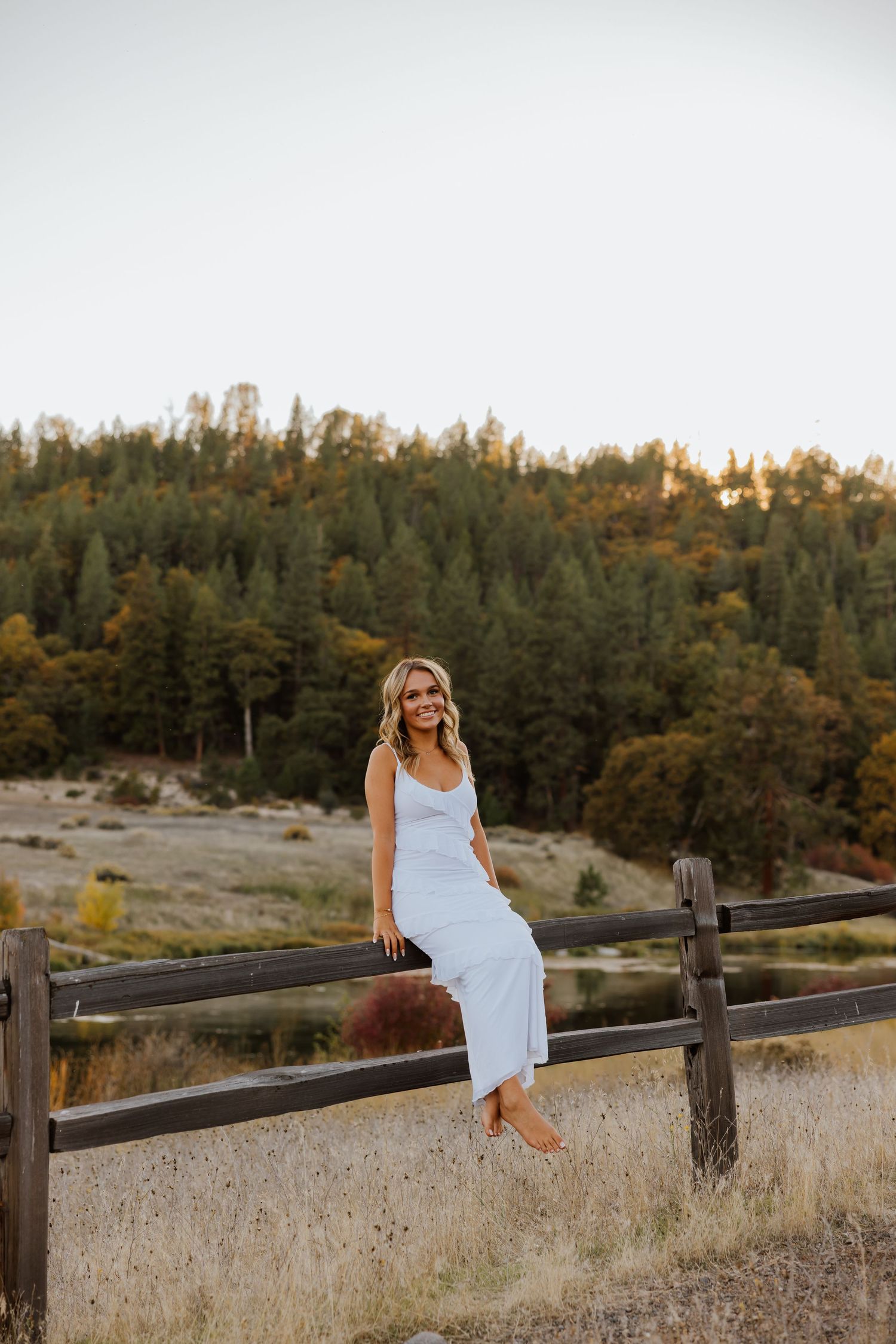 Woman in white dress sits on wooden fence with forested hillside in background.