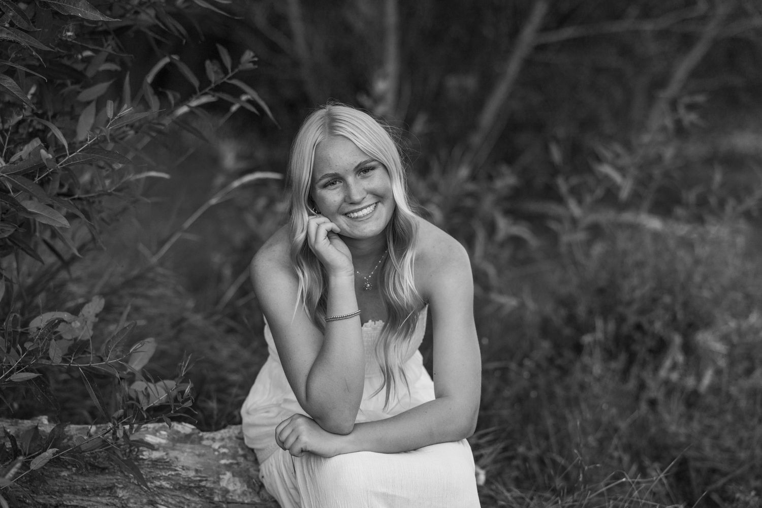 Black and white portrait of someone in white dress sitting outdoors with natural foliage background.