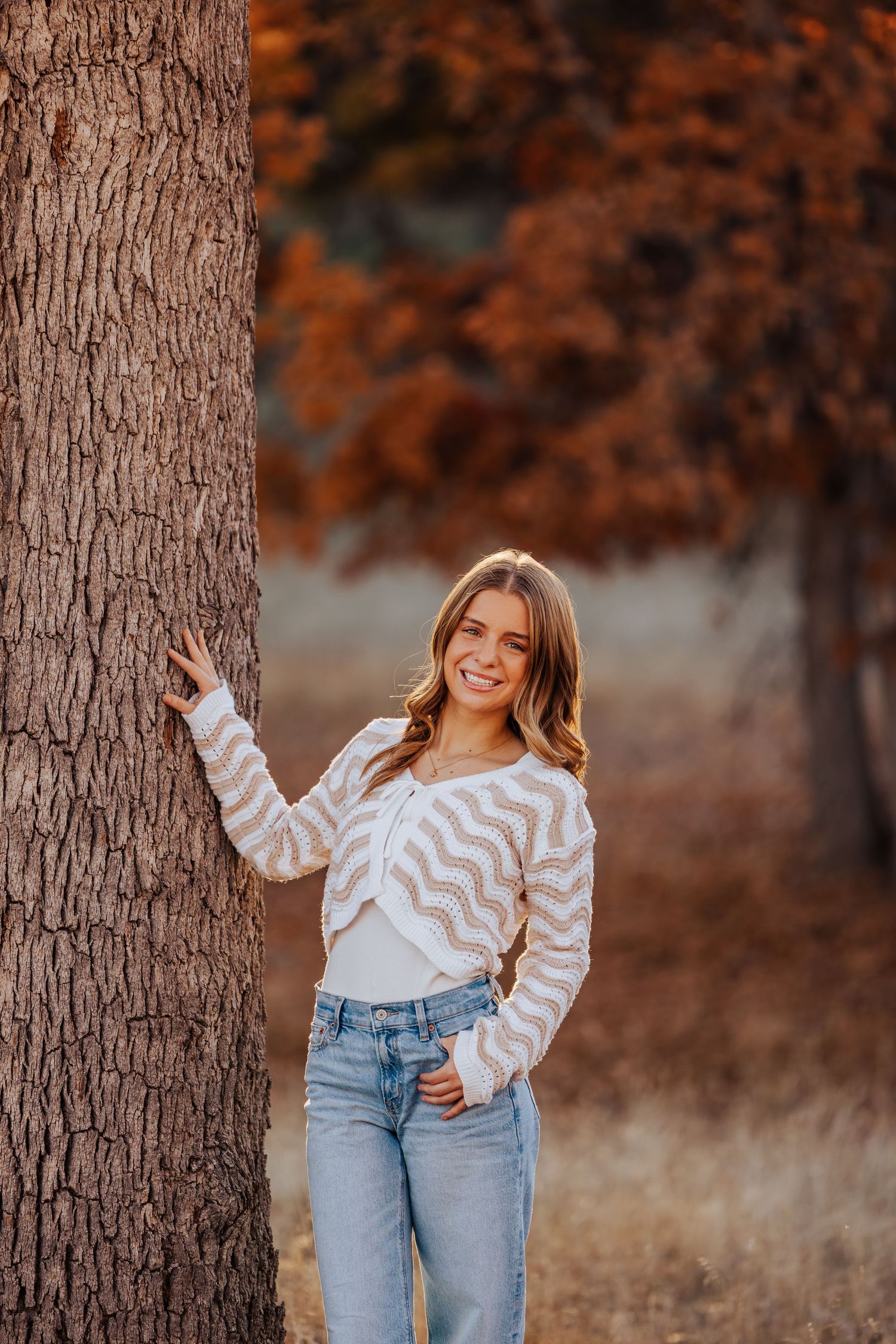 Portrait against tree trunks wearing a chevron sweater and jeans in warm autumn lighting.