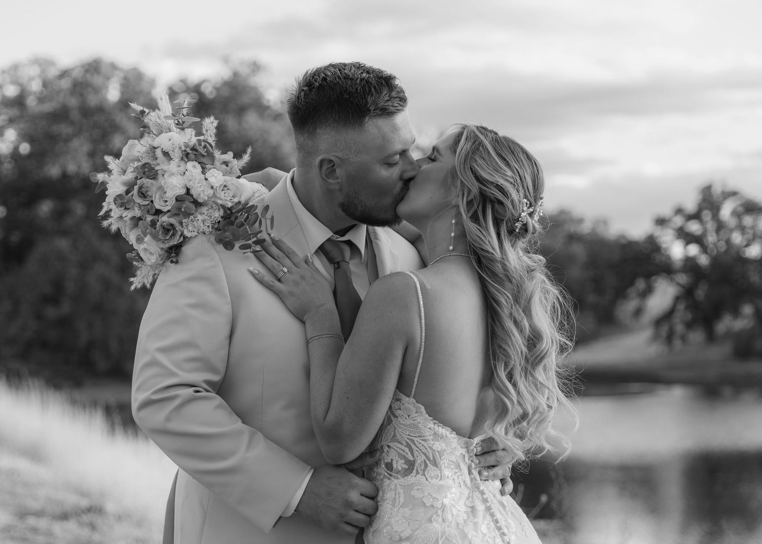 A romantic black and white wedding photo of a couple kissing while holding a floral bouquet outdoors.