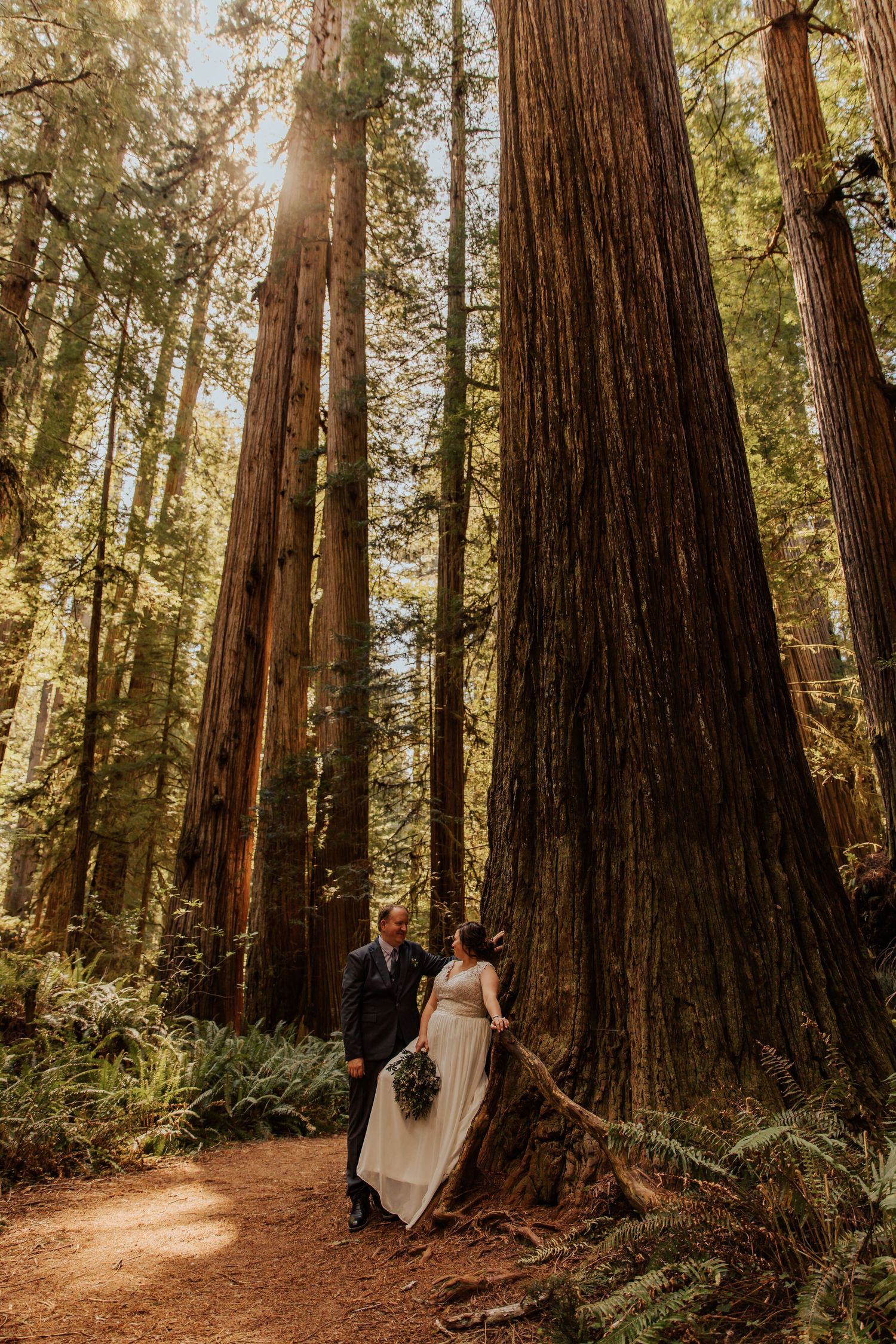 A couple shares a romantic moment on a sunlit forest path surrounded by towering redwood trees.