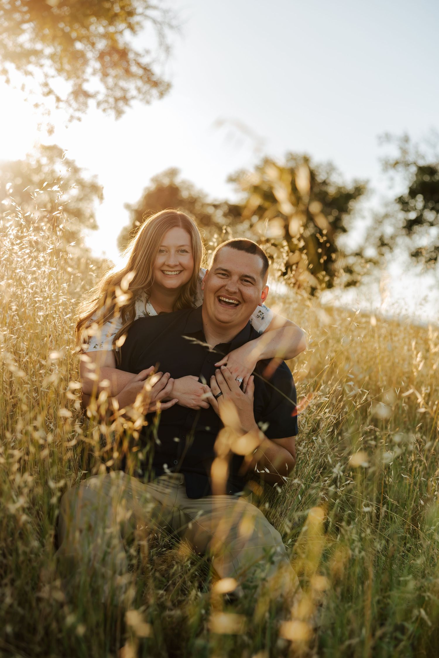 Couple laughing together in golden sunlit field with tall grass and wildflowers during sunset engagement session.