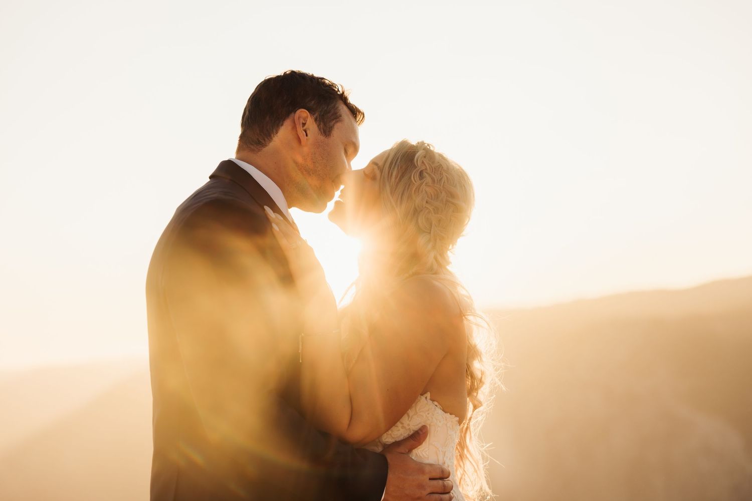 Couple sharing a romantic kiss at golden hour with warm sunlight glowing behind them in an outdoor wedding photo.
