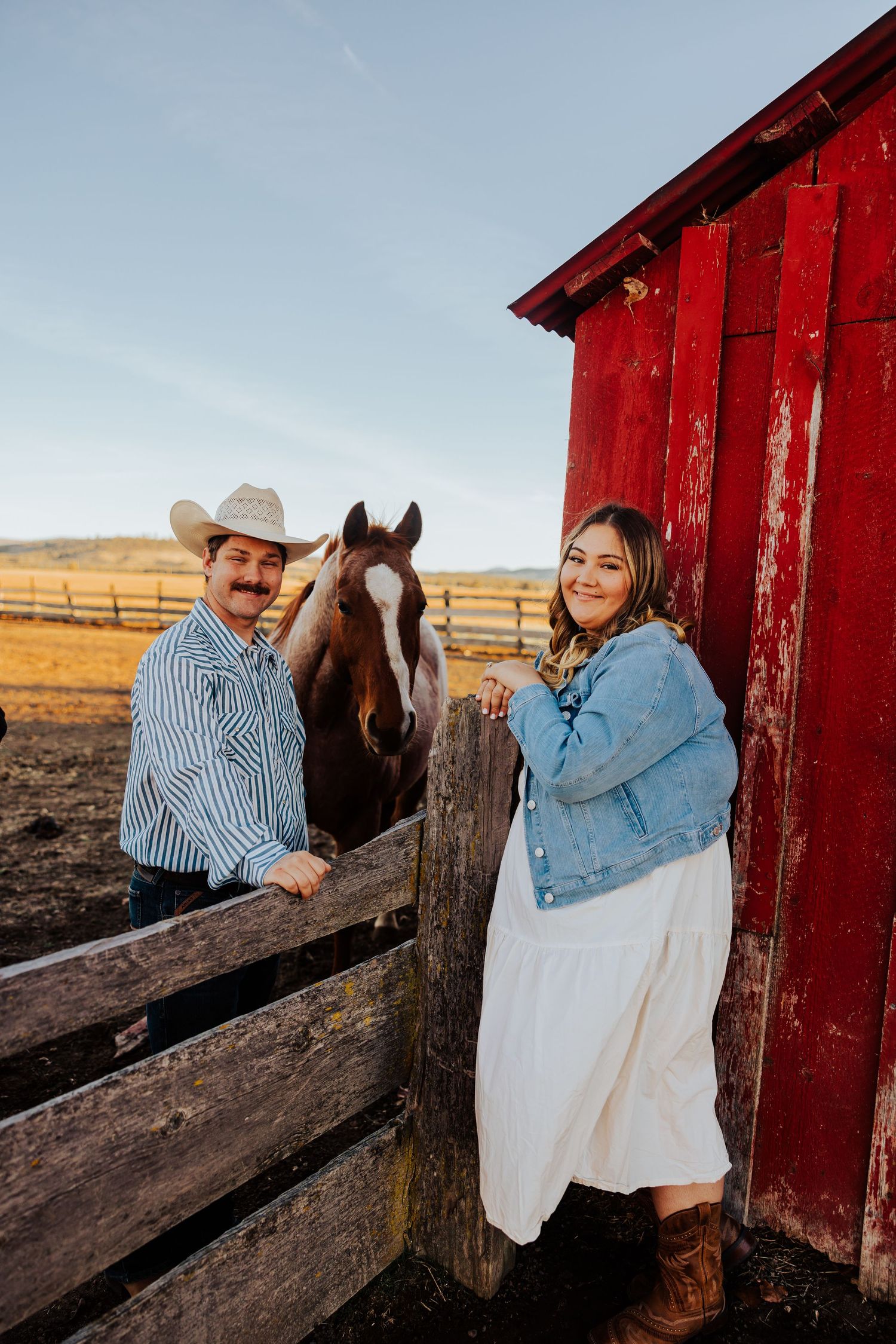 A couple poses near a red barn and wooden fence with a horse looking on.