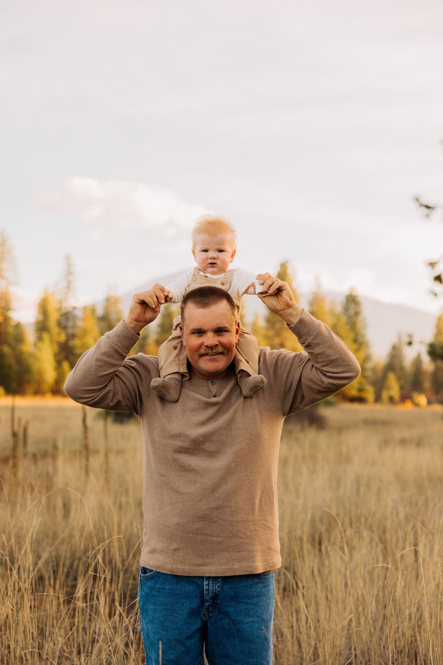 A playful father lifts his baby overhead in a mountain meadow during golden hour.