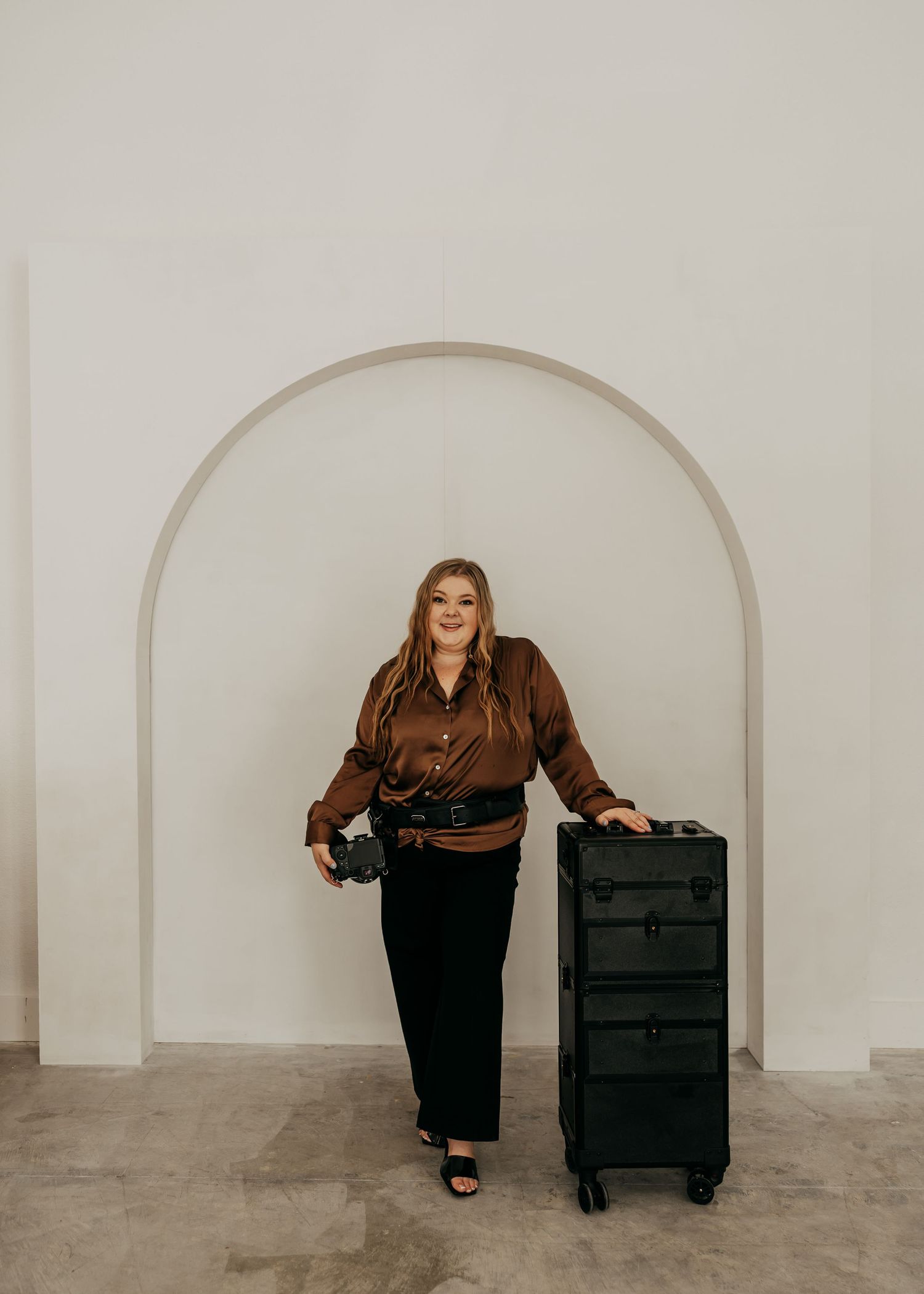 Woman in brown satin blouse poses beside black rolling makeup case in front of white arched wall.