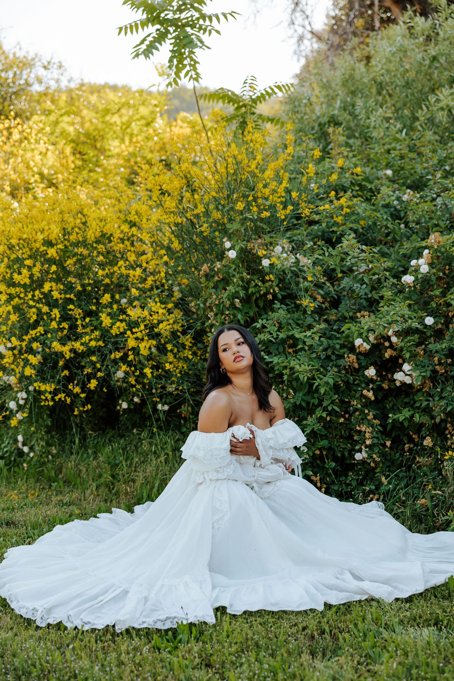 Bride in white off-shoulder gown sitting on grass surrounded by yellow and white flowering bushes outdoors.