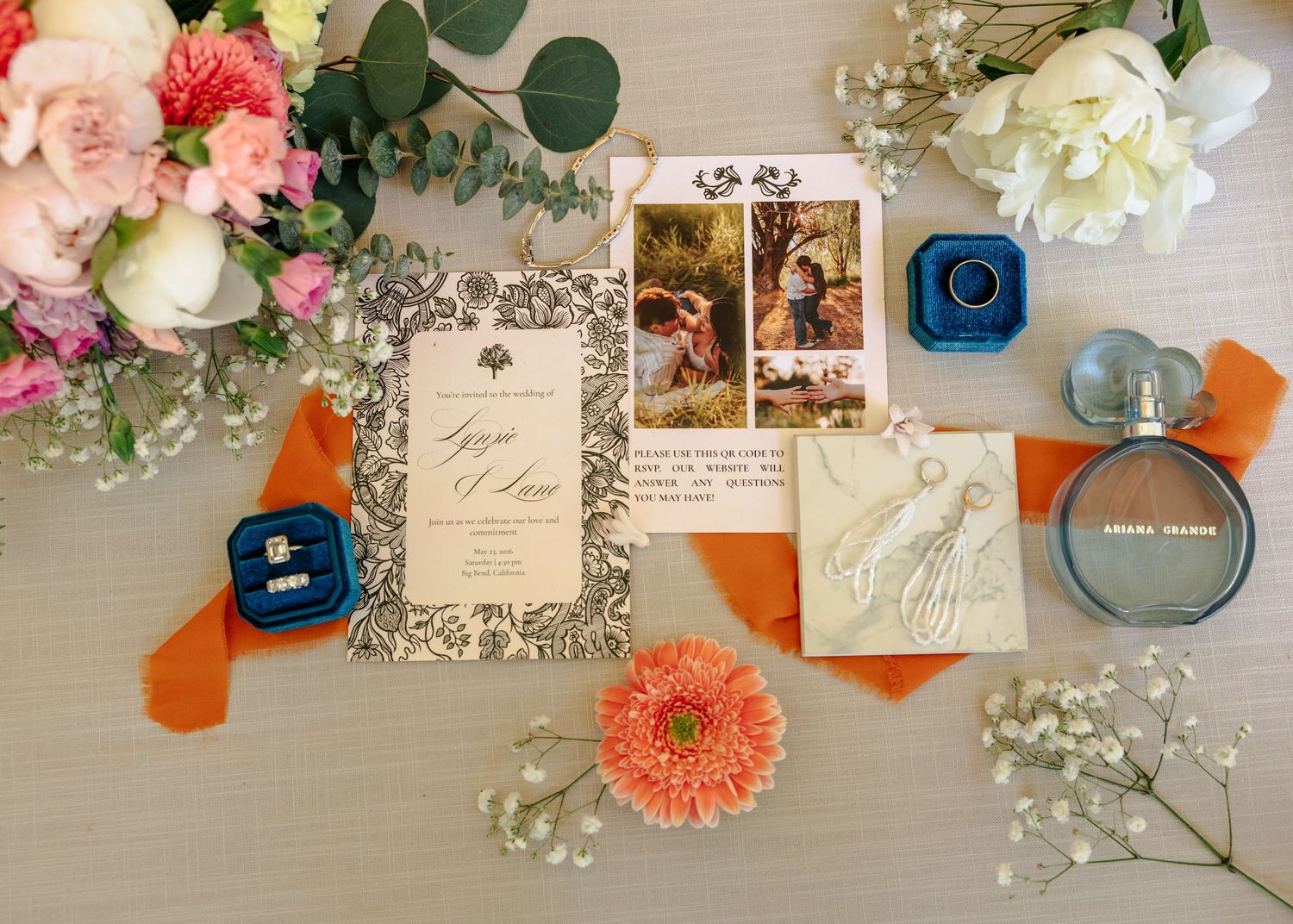 Elegant wedding flat lay with floral invitations, orange ribbon, ring boxes, perfume bottle, and pink flowers on white background.