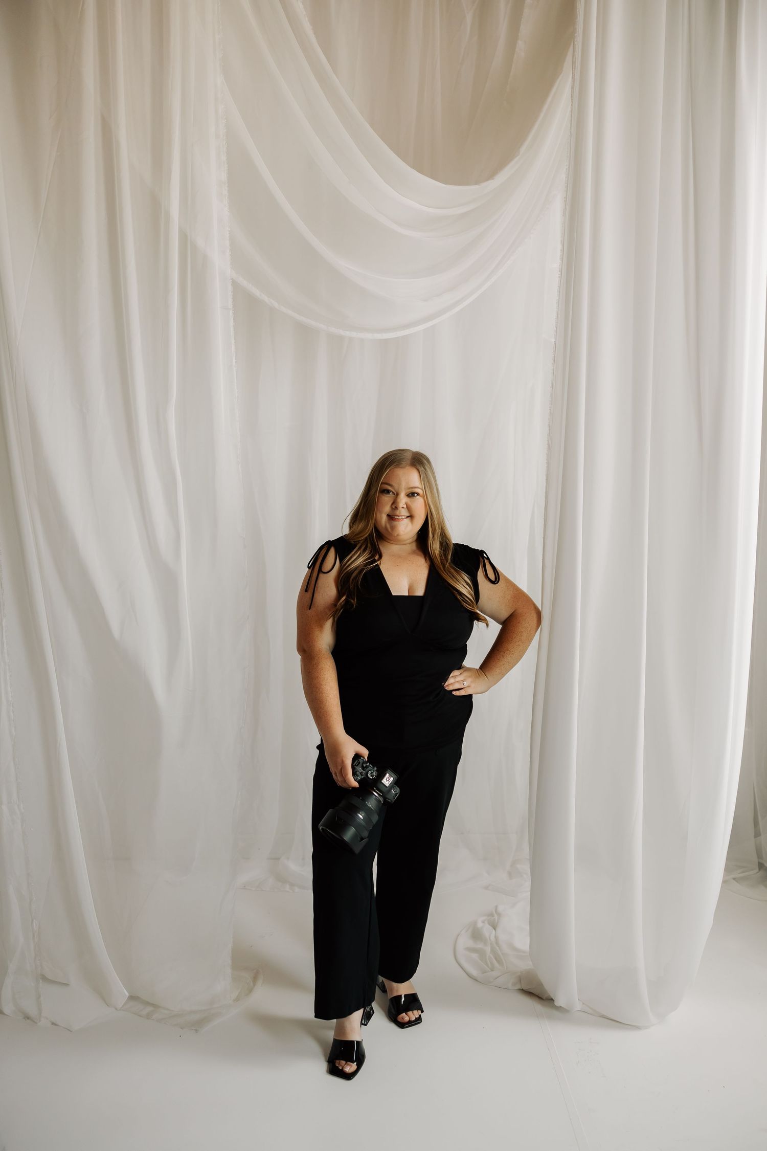 Woman in black jumpsuit holding camera, smiling in front of white draped fabric backdrop.