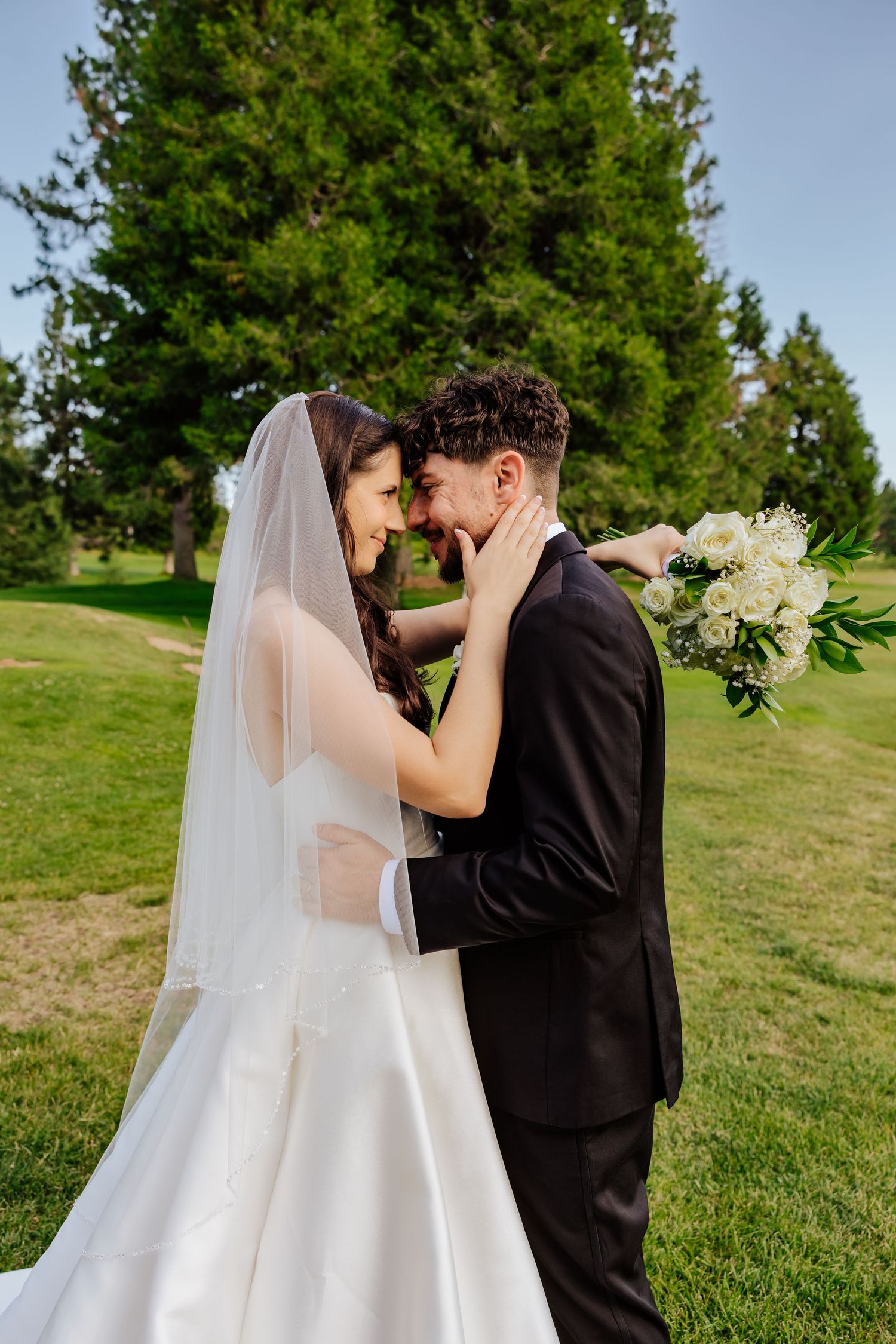 Bride and groom share a romantic moment outdoors, touching foreheads, as bride holds white bouquet in a lush green park.