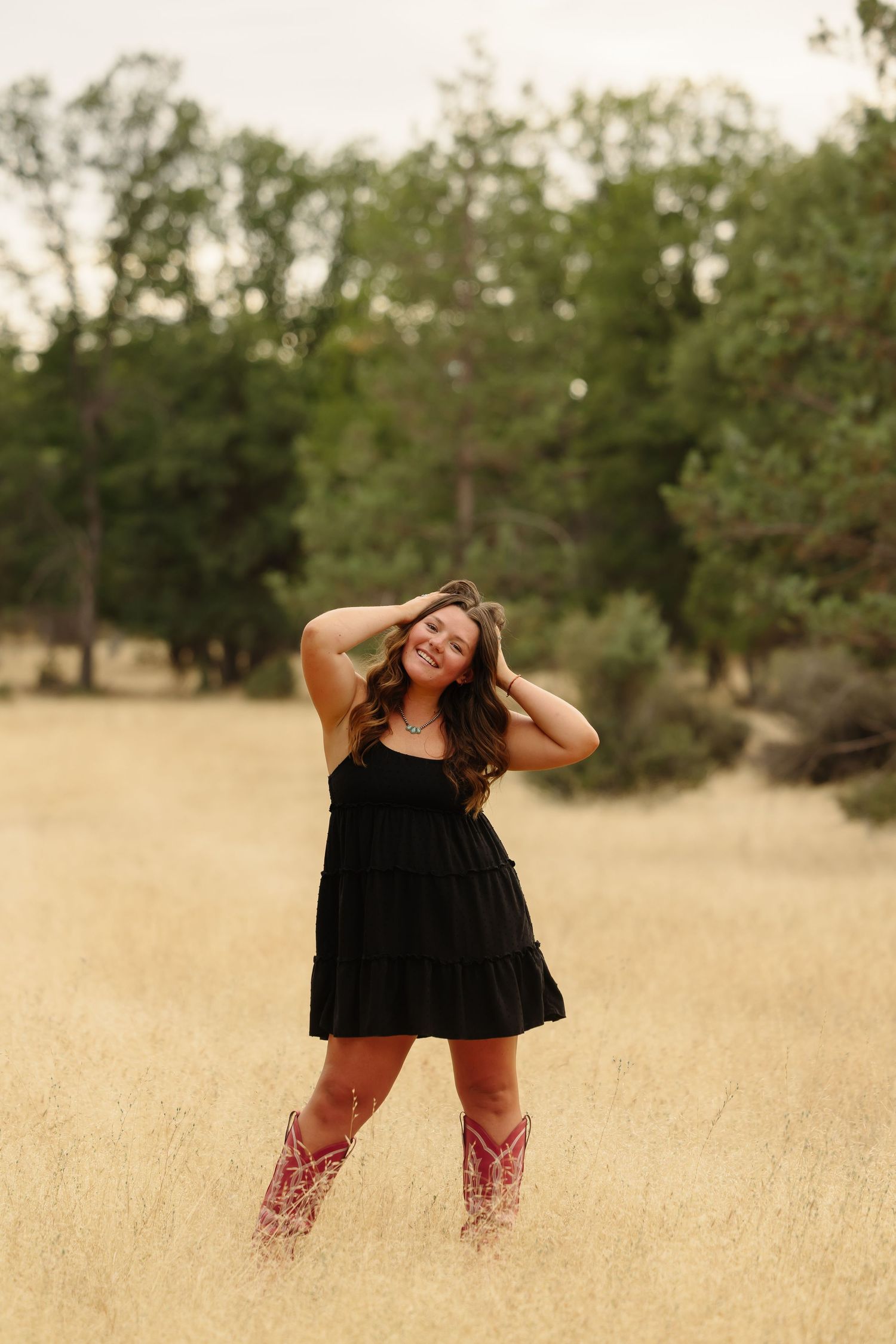 Young woman in black mini dress and red cowboy boots smiling in golden field with trees behind her.