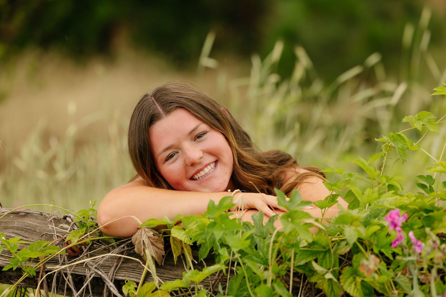 Smiling young woman with long brown hair resting on a log surrounded by lush green plants and wildflowers outdoors.